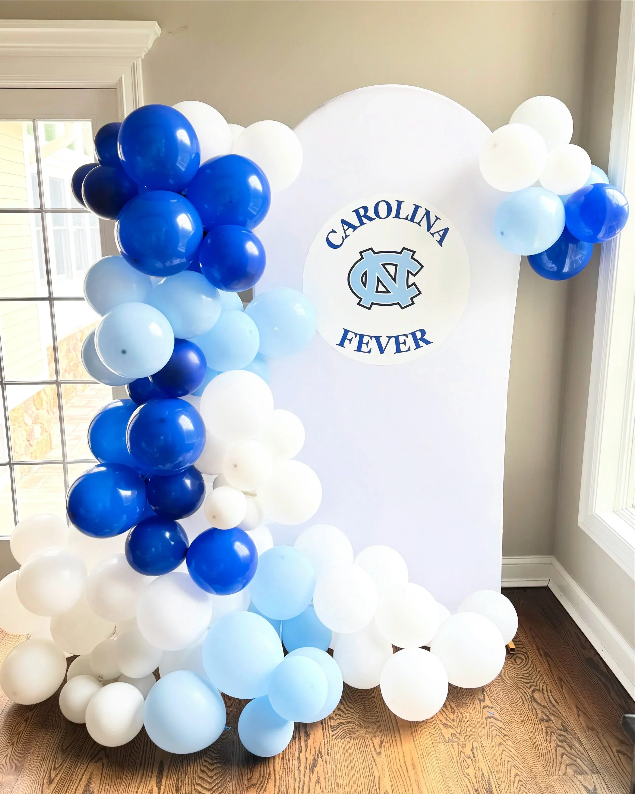 Balloon arrangement with white, light blue, dark blue, and black balloons positioned in front of a white backdrop with a circular emblem that has the North Carolina Tar Heels logo and the words "Carolina Fever."