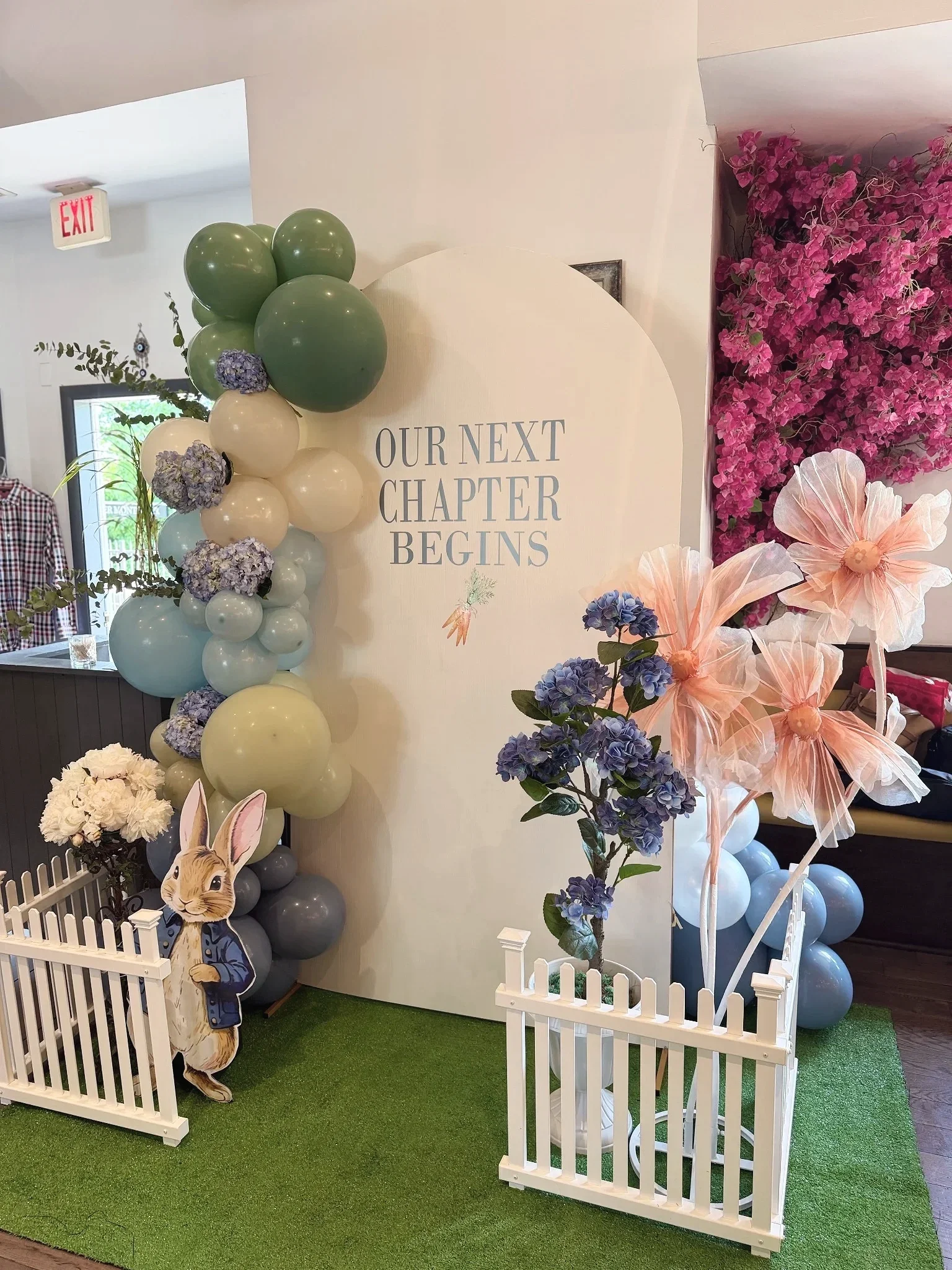 Decorative baby shower display with pastel balloons, pink flowers, a bunny cutout, and a sign that reads "Our Next Chapter Begins" for a celebration or event.