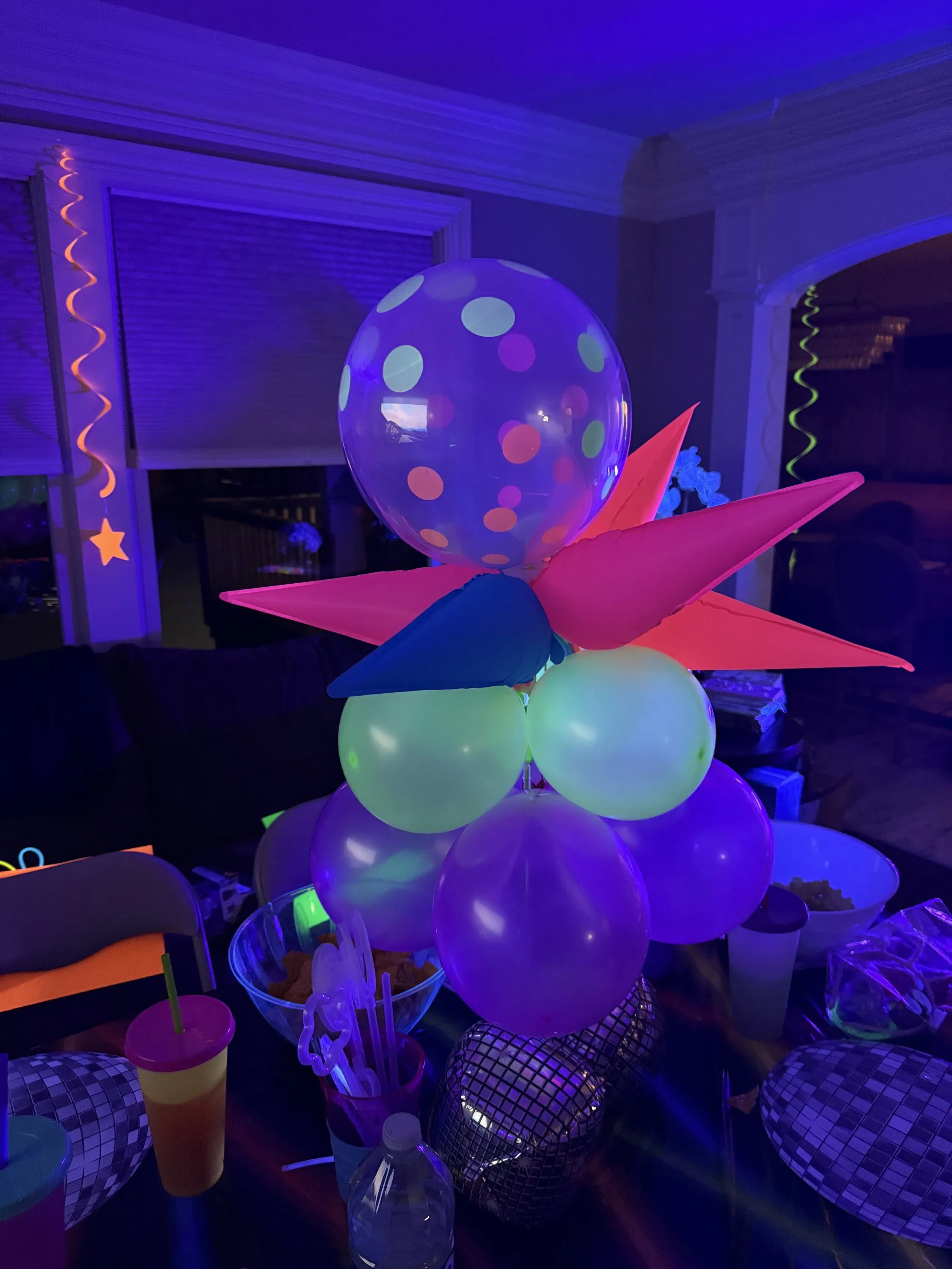 Colorful balloons, paper decorations, and party supplies illuminated by black lights at a celebration or party.