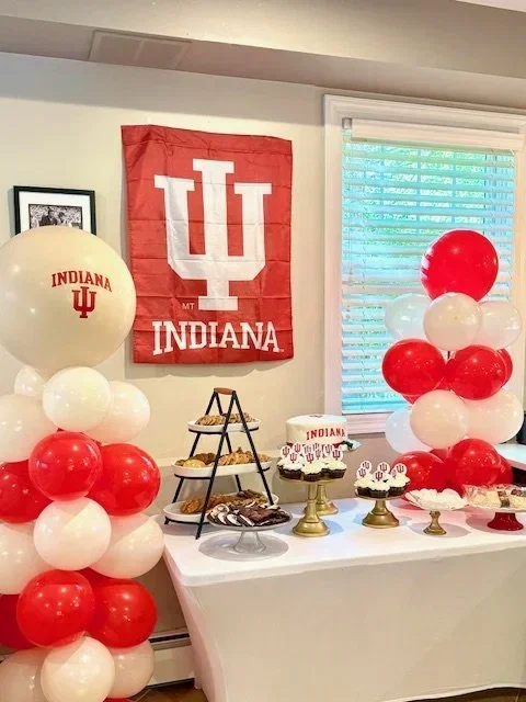 Decorative table with Indiana University themed balloons, cakes, and snacks in a room with an Indiana flag on the wall and a window with blinds.