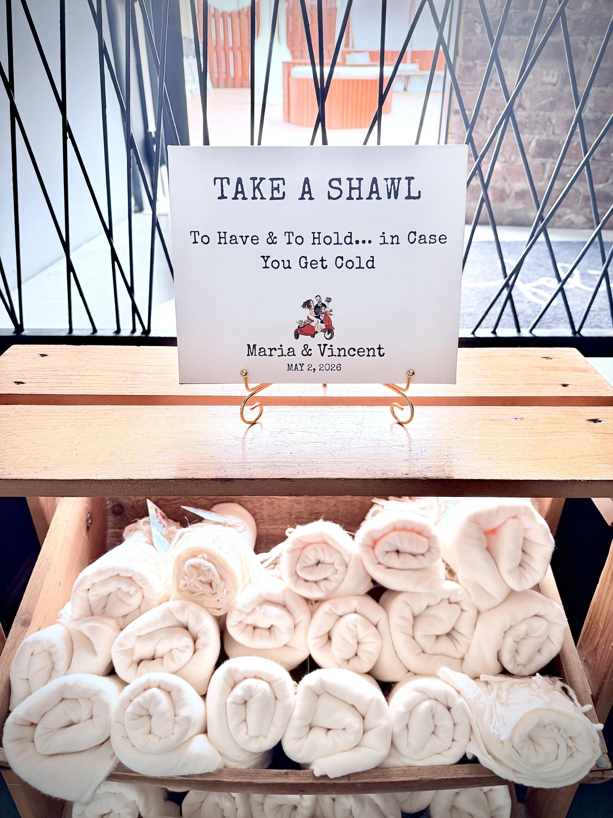 A display of neatly rolled white towels on a wooden shelf with a sign that reads "Take a Shawl To Have & To Hold... in Case You Get Cold" and decorative black metal bars behind it.