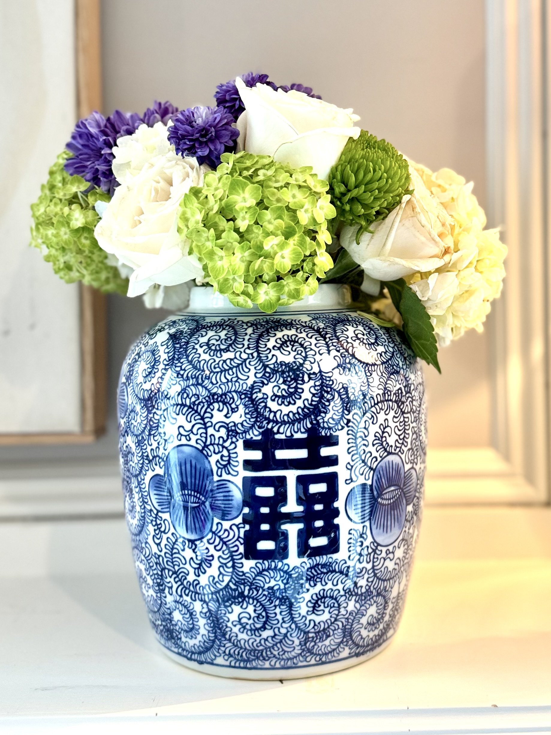 A decorative blue and white floral-patterned vase filled with white roses, purple flowers, green hydrangeas, and cream-colored roses.