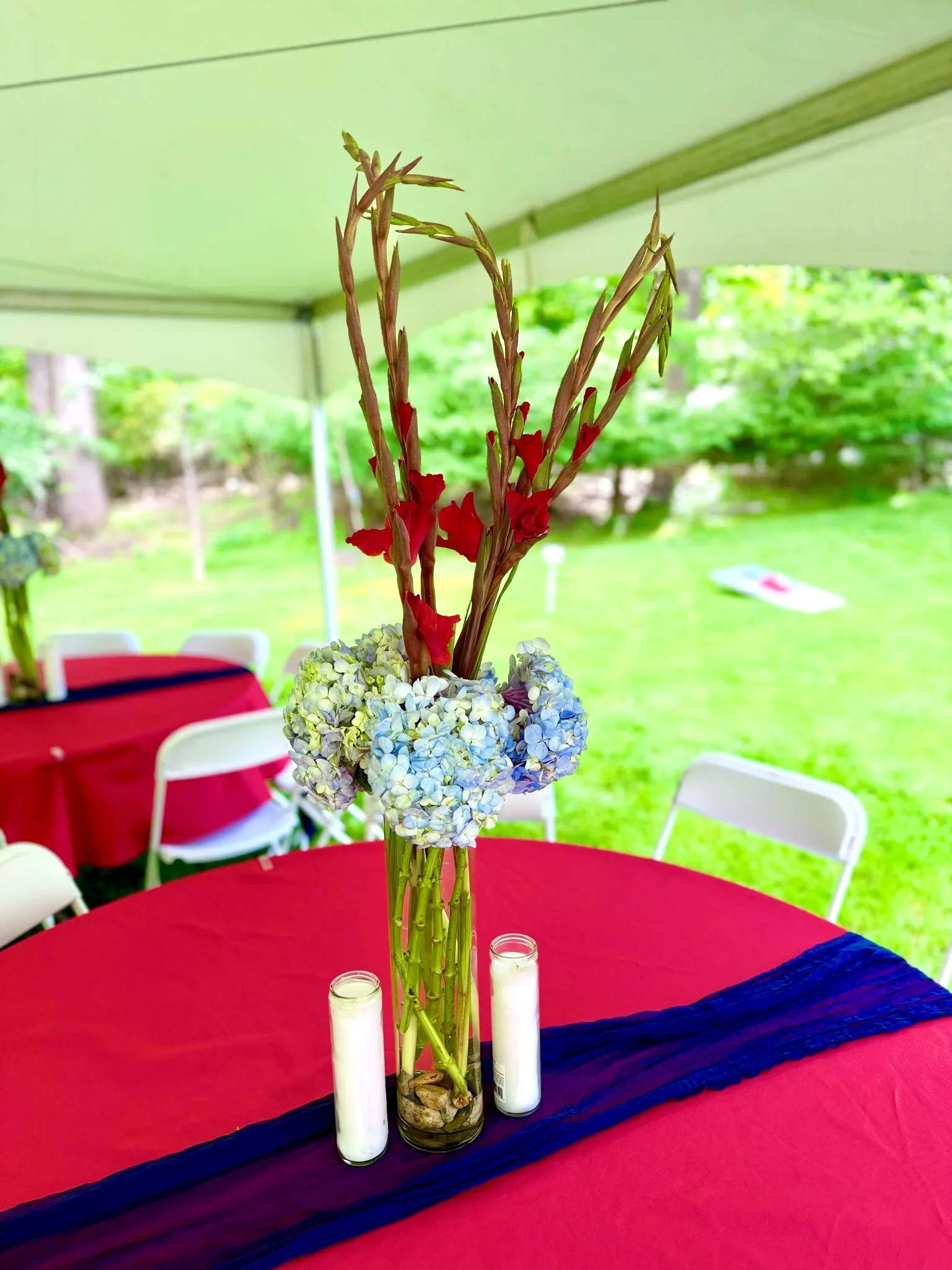 Centerpiece with tall flower arrangement of red gladiolus and white hydrangeas on a red table, decorated with white candles and a navy blue runner, outdoors at daytime.
