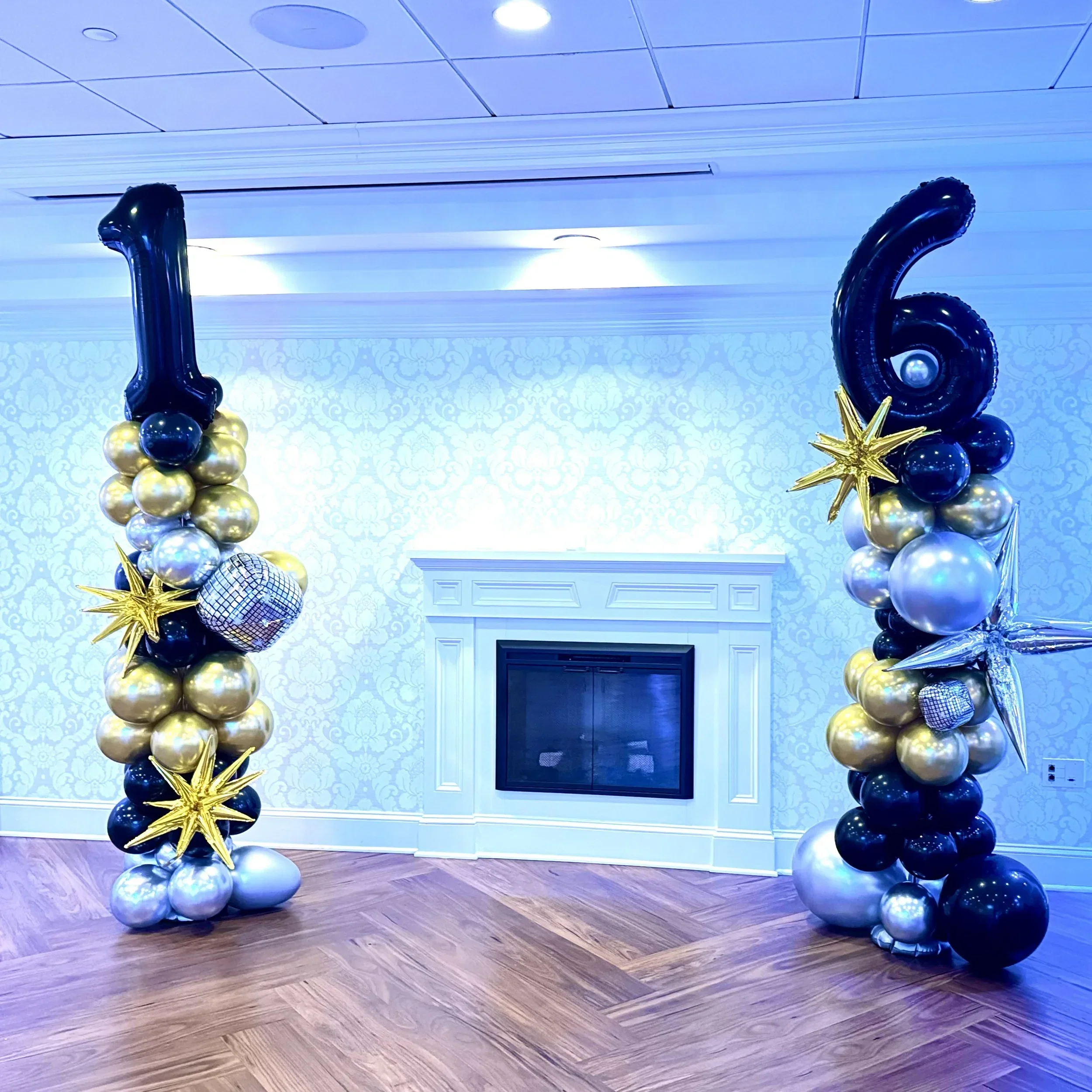 Balloon arch with black, gold, and silver balloons, shaped as numbers 1 and 6, with gold star balloons and a glittery disco ball, set against a decorated wall and fireplace.