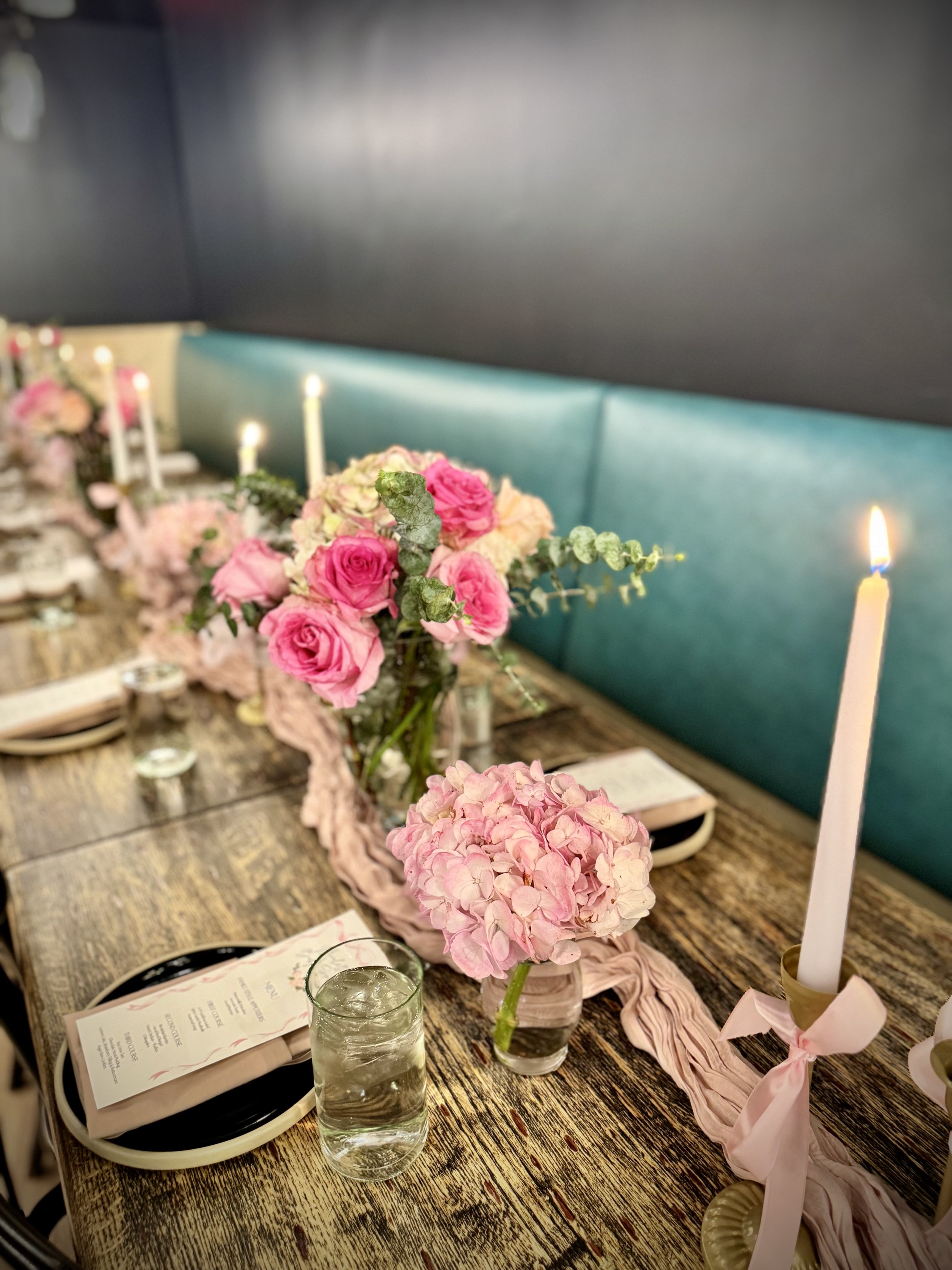 A decorated dining table with pink and white flowers in glass vases, lit candles, and place settings with menus and glasses, set for a celebration or event.