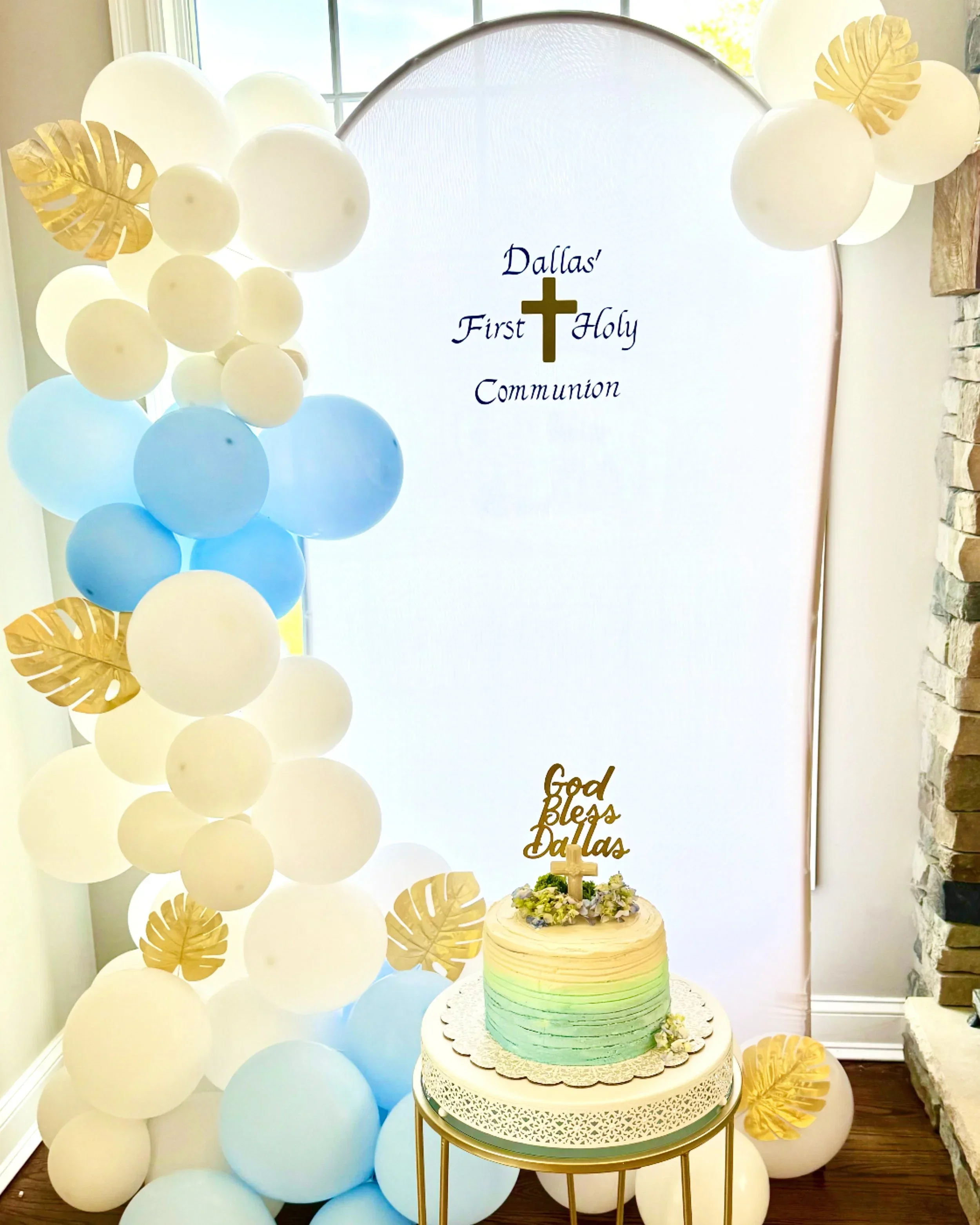 Decorative setup for a First Holy Communion celebration, featuring a large white backdrop with the text "Dallas' First Holy Communion" and a gold cross, surrounded by white and pastel balloons, gold leaf accents, and a cake with a cross and floral to