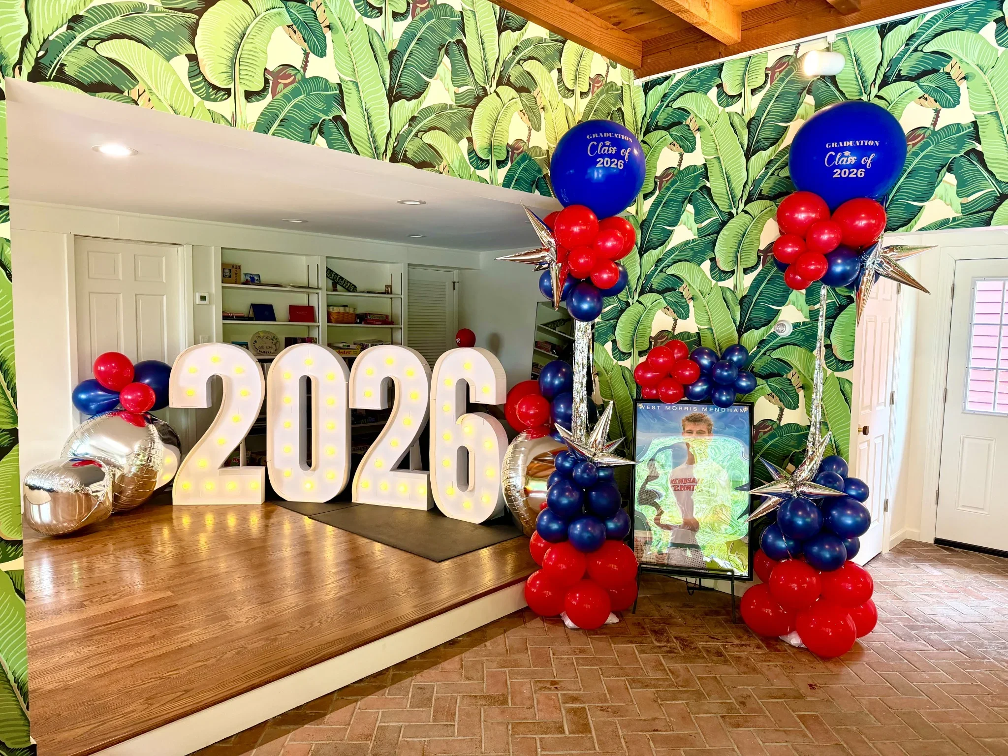 Decorations for a graduation celebration, featuring large illuminated numbers '2026,' red, blue, and silver balloons, and green leafy wallpaper.
