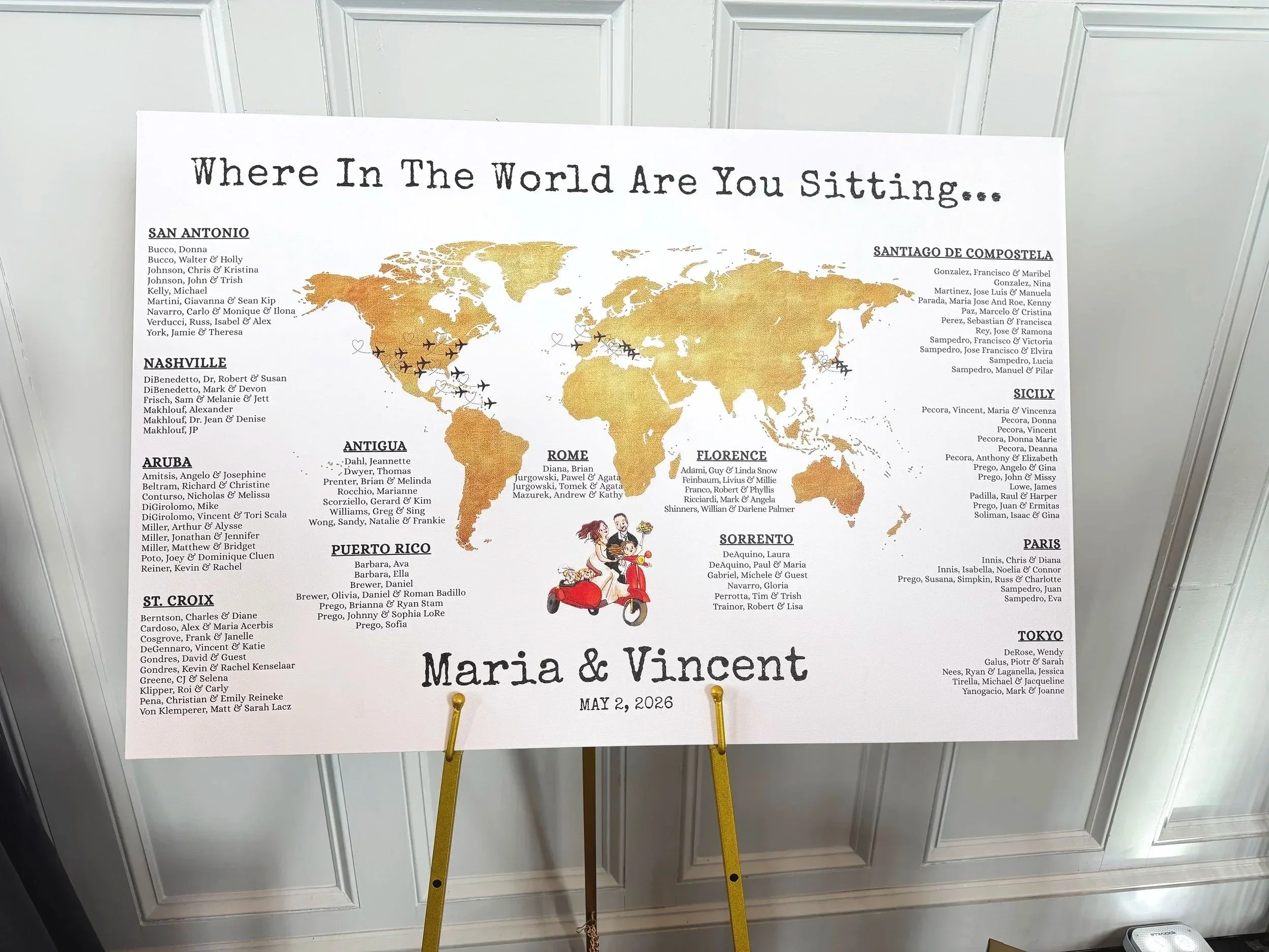 A seating chart or sign with the title 'Where In The World Are You Sitting...' and the names of different locations such as San Antonio, Nashville, Antigua, Puerto Rico, St. Croix, Sorrento, Florence, Sicily, Paris, and Tokyo, along with corresponding guest names for each location. The sign also features a world map illustration and the names 'Maria & Vincent' with the date 'May 2, 2026' at the bottom.