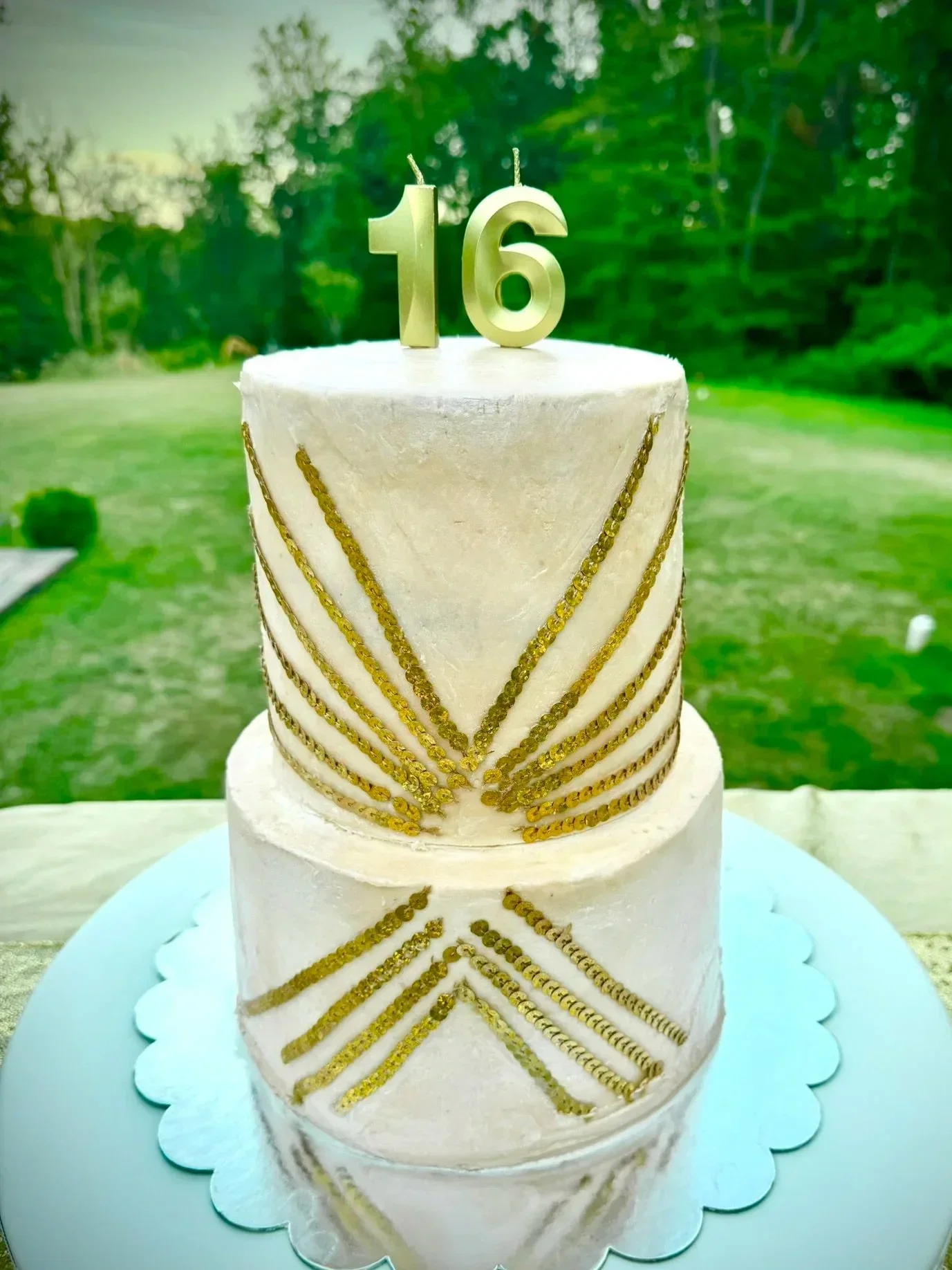 A two-tiered white cake decorated with gold chains and beads, with gold number candles displaying '16' on top, set outdoors on a light blue cake stand.