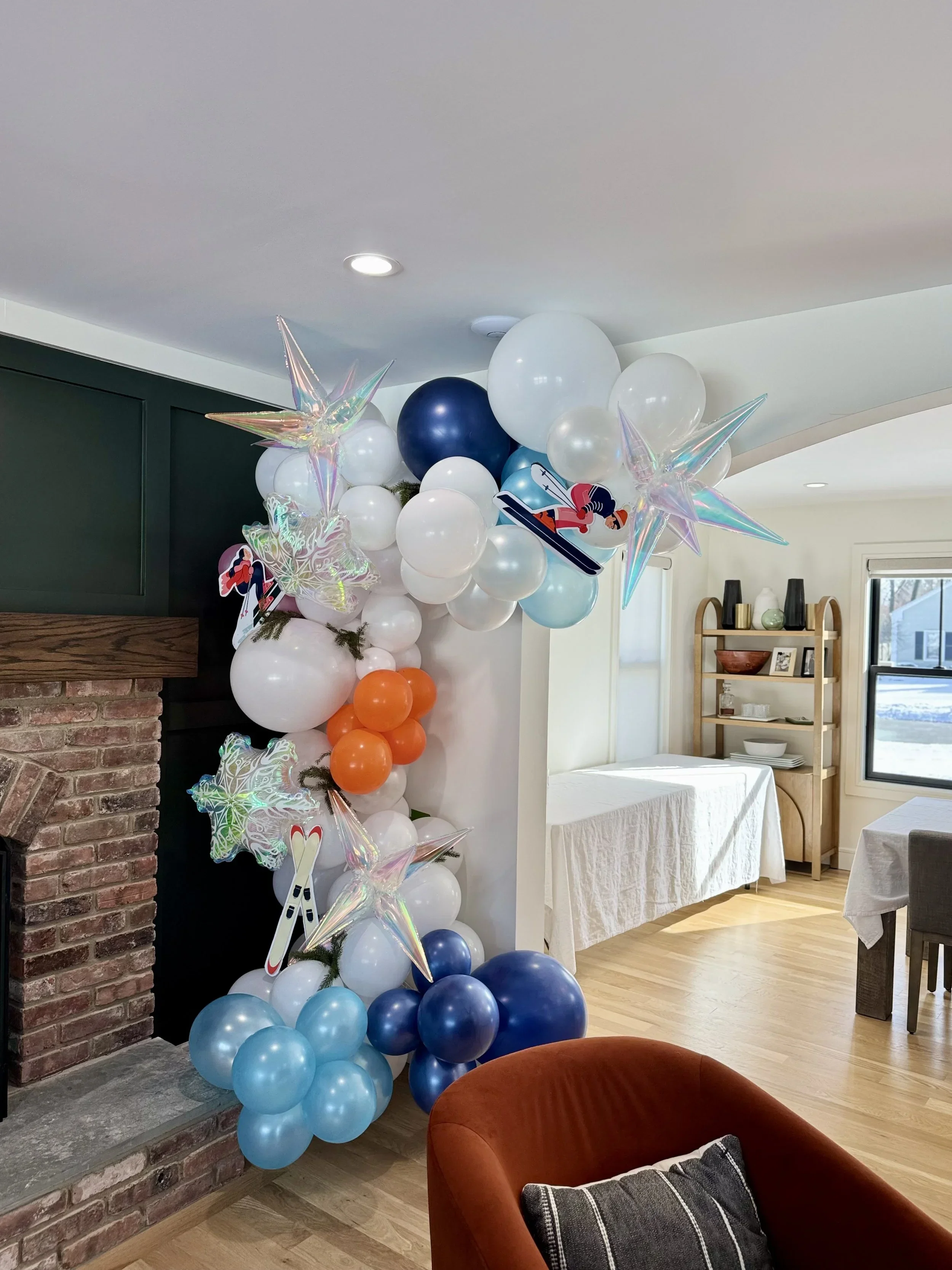 Indoor party decoration with a balloon arch featuring white, blue, and orange balloons, holographic star-shaped balloons, and cartoon character cutouts in a living room with a brick fireplace, bookshelf, and dining area.