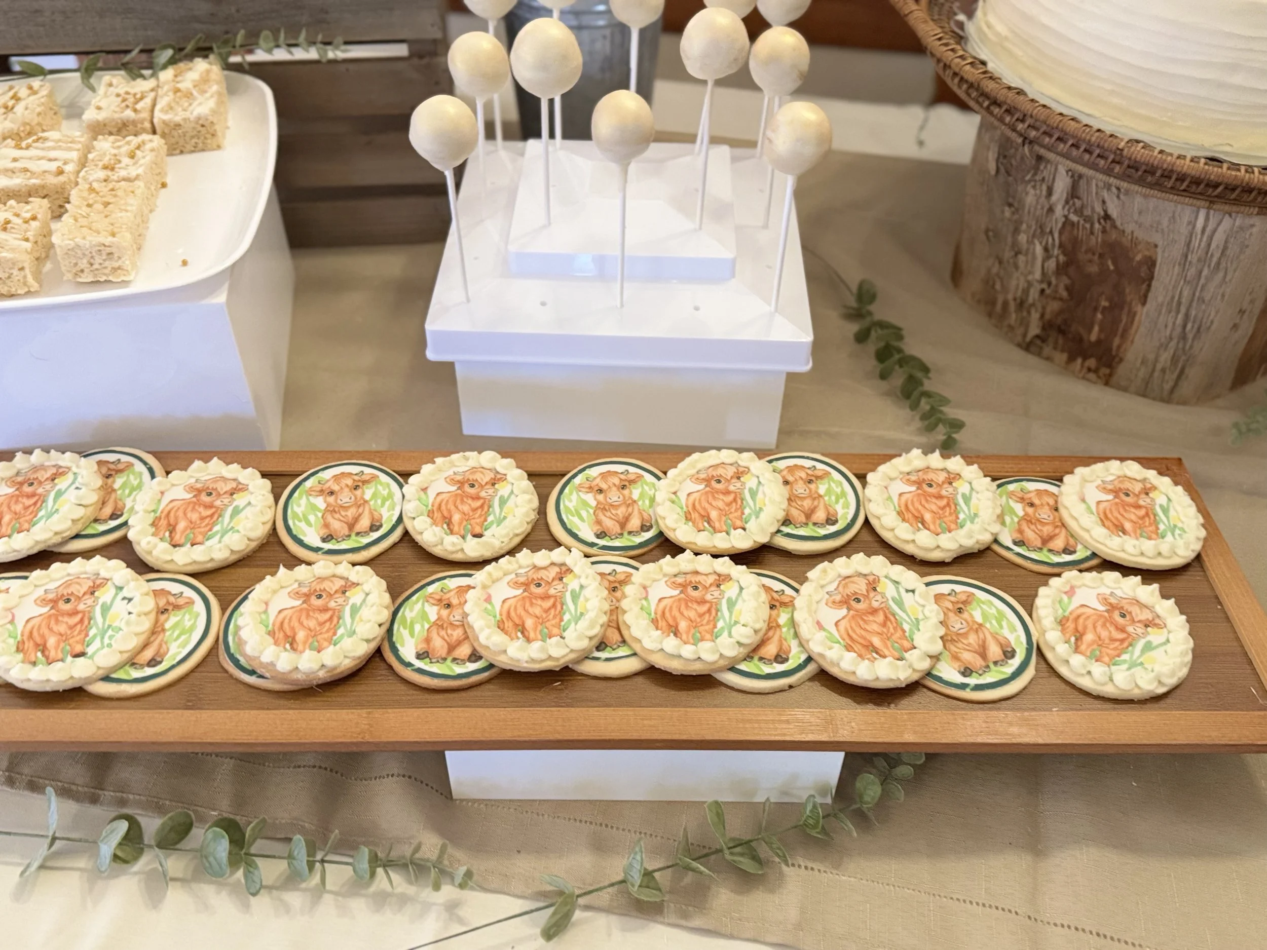 Cookies with cow-themed icing on a wooden serving tray surrounded by other desserts.