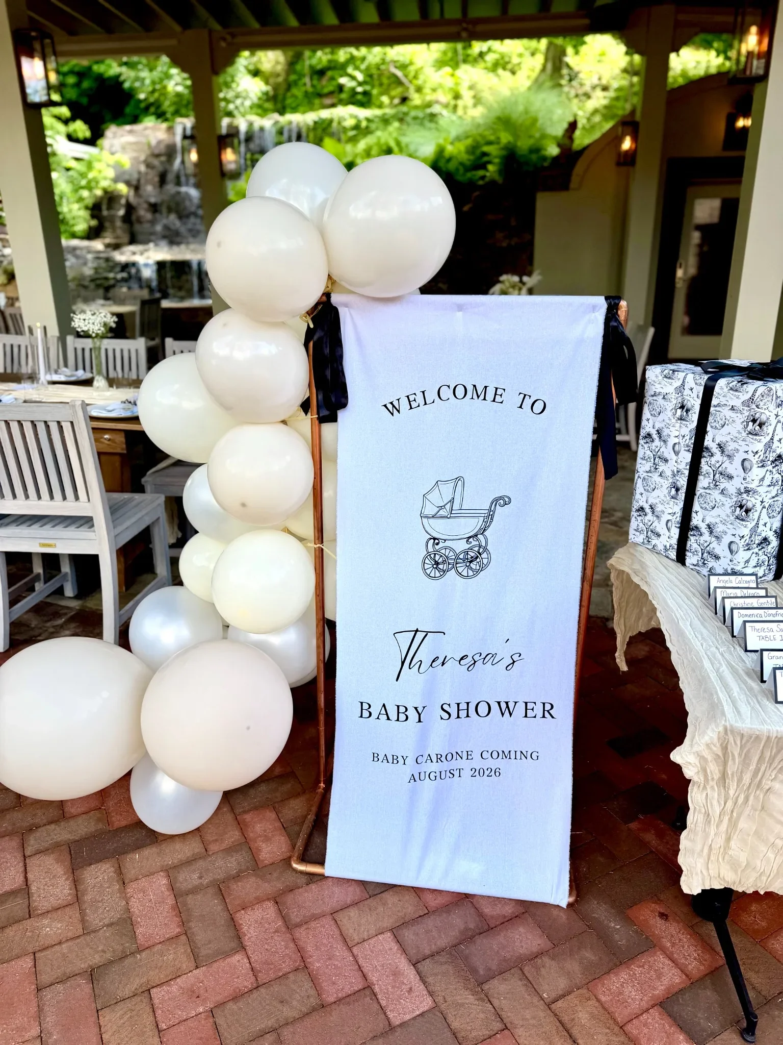 Decorative baby shower sign with white balloons attached, welcoming guests to Theresa's baby shower scheduled for August 2026, with a scenic outdoor background including green trees and a waterfall.
