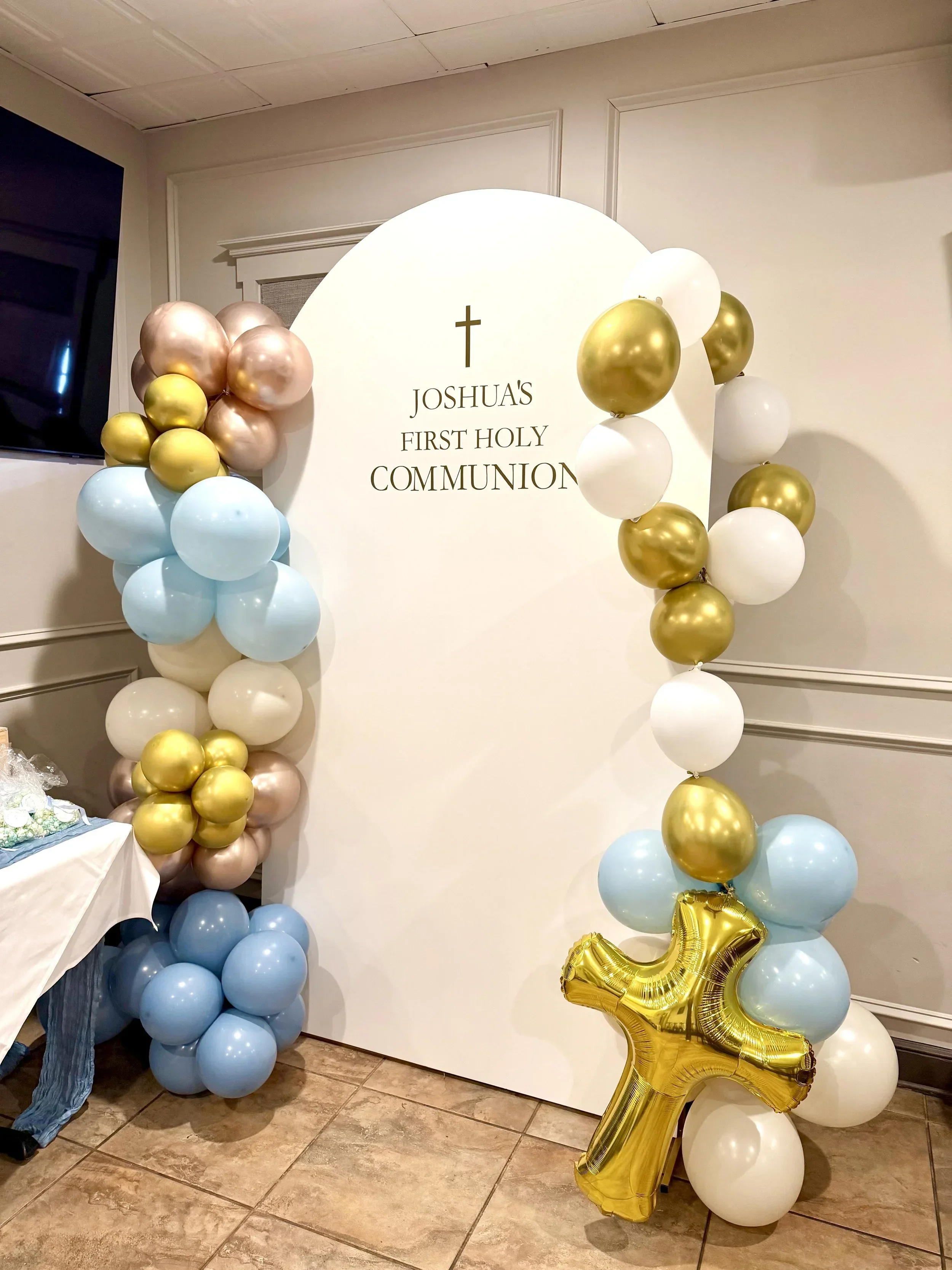 A white backdrop with black text reading "Joshua's First Holy Communion," decorated with pastel-colored balloons in gold, white, light blue, and pink, arranged on both sides and at the bottom, with a large gold number '1' balloon at the bottom right.