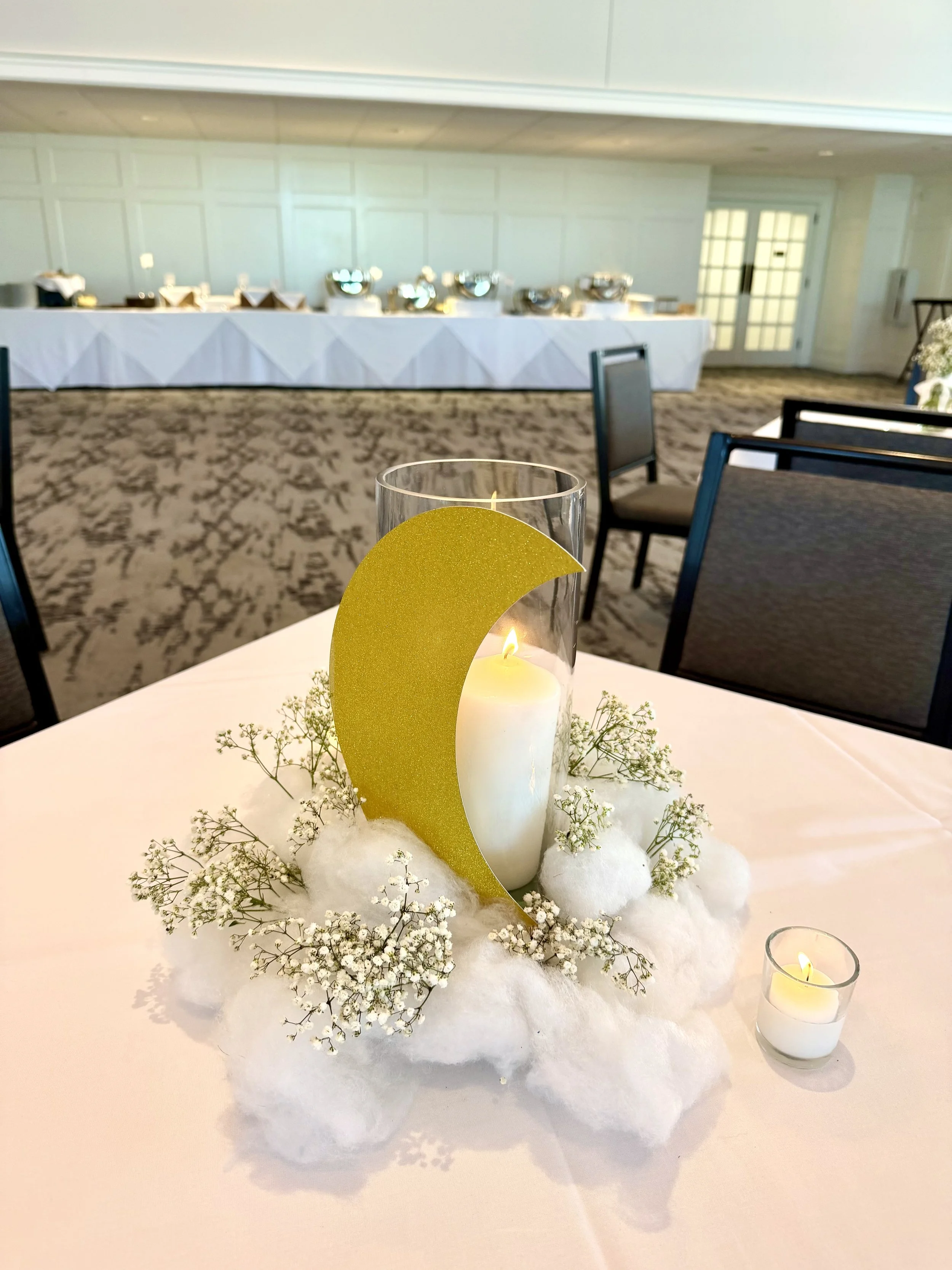 Decorative table centerpiece with white candles, gold crescent moon, white cotton balls, baby's breath flowers, and cotton-like cotton balls, with small votive candles, on a white tablecloth at an event venue in the background.