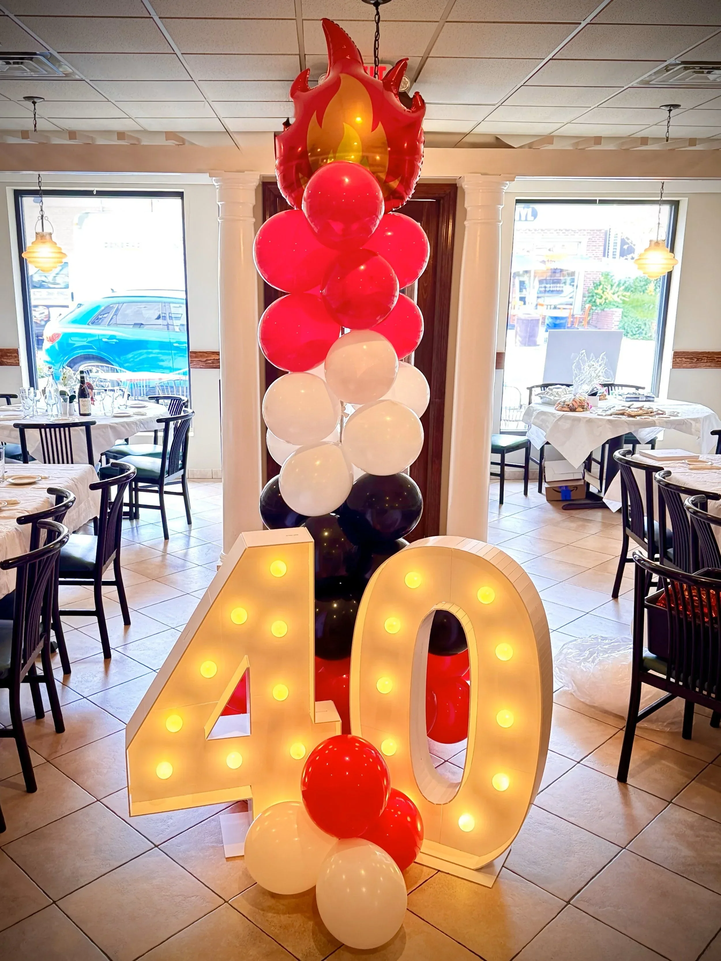 Decorative fire-themed balloon arrangement with a large flame-shaped balloon, red and white balloons, and a light-up 'LOVE' sign in a restaurant or event space.