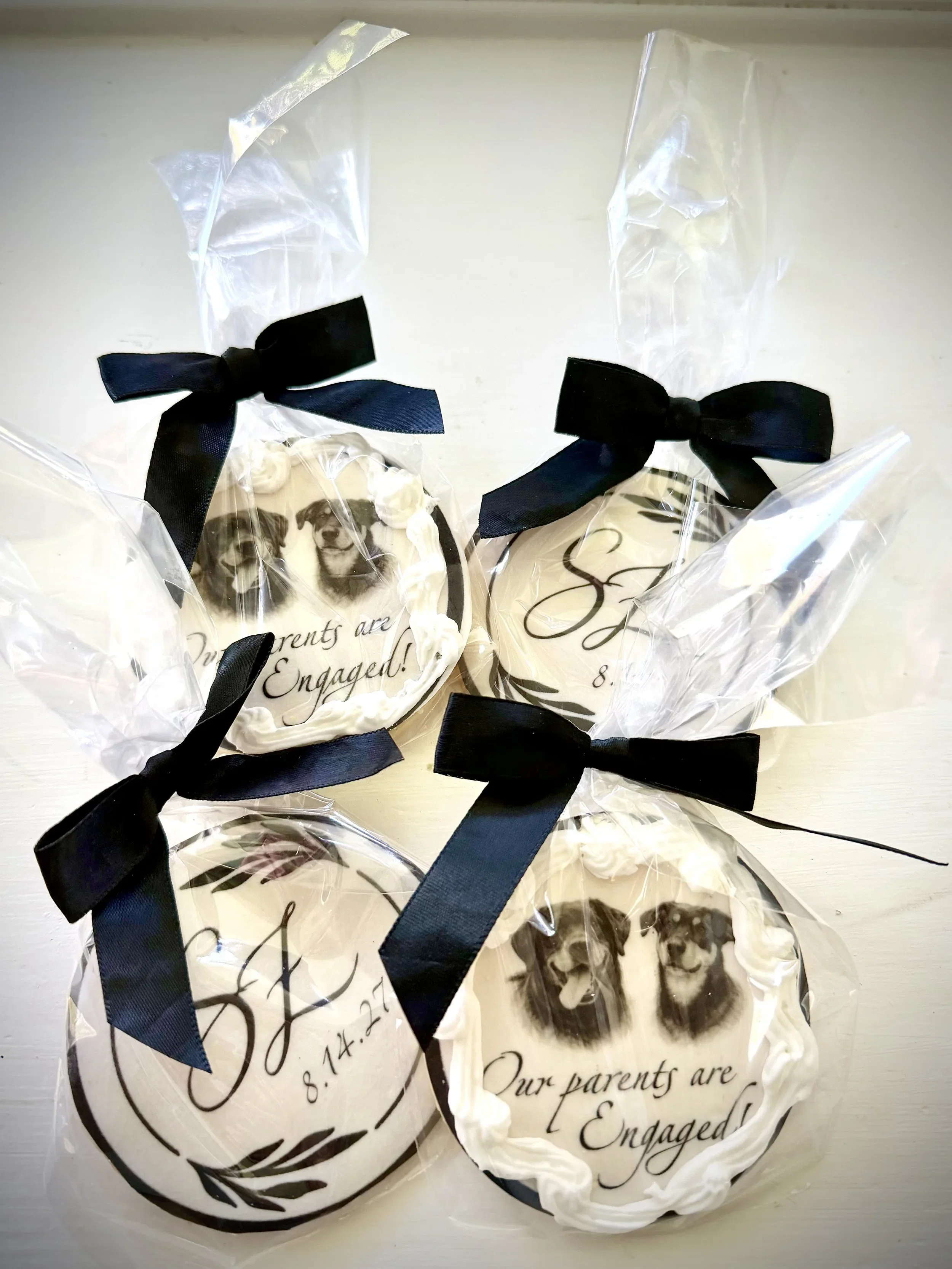 Two decorated cookies wrapped in clear plastic with black ribbons, one with the word Engagement and a photo of a couple, the other with the date August 27 and the phrase Our parents are engaged.