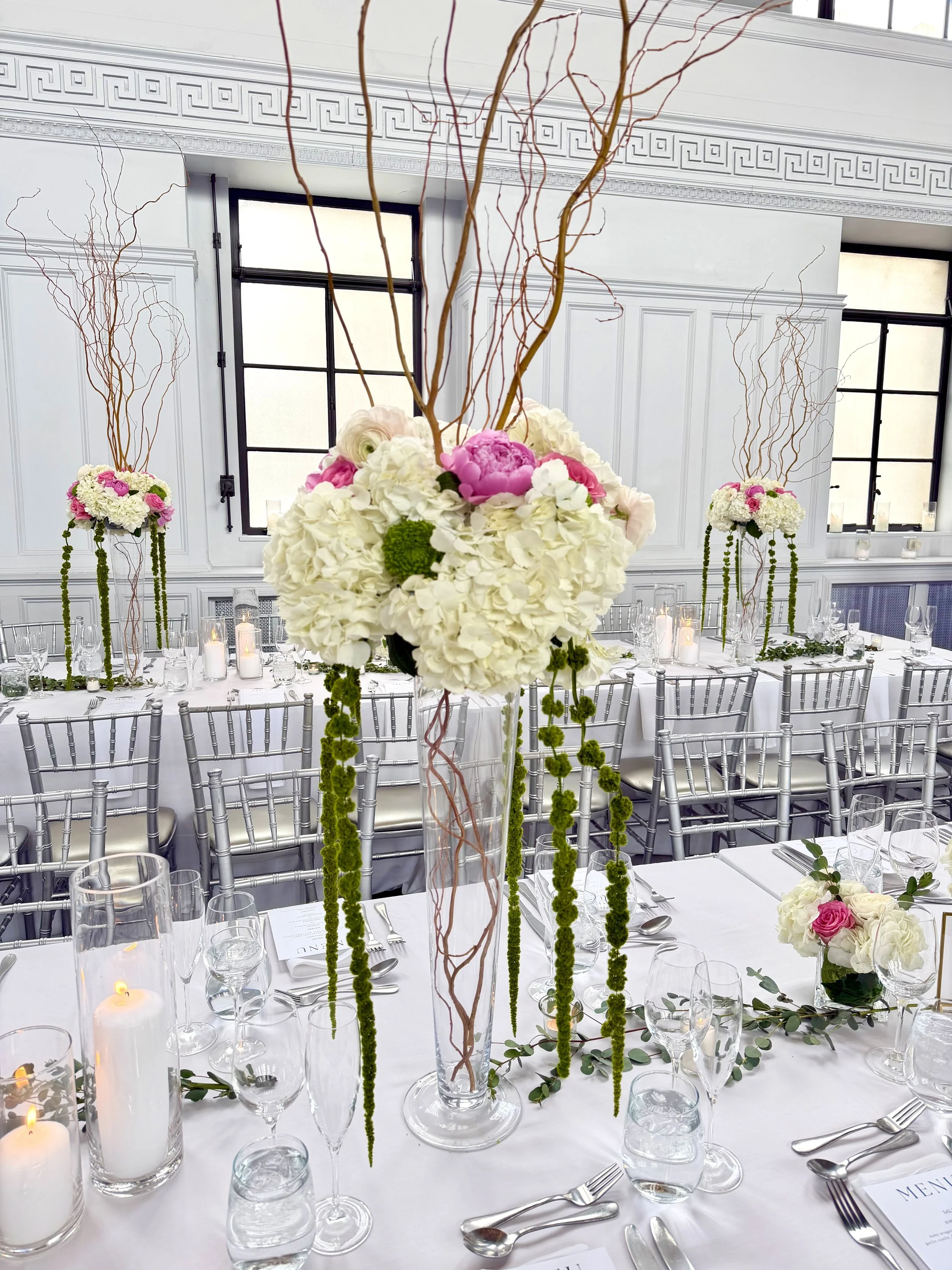 Elegant dining table setting with white tablecloth, floral centerpieces with white and pink flowers, tall glass vases with green hanging accents, candles, glasses, silverware, and place cards in a bright room with large windows and white ornate walls