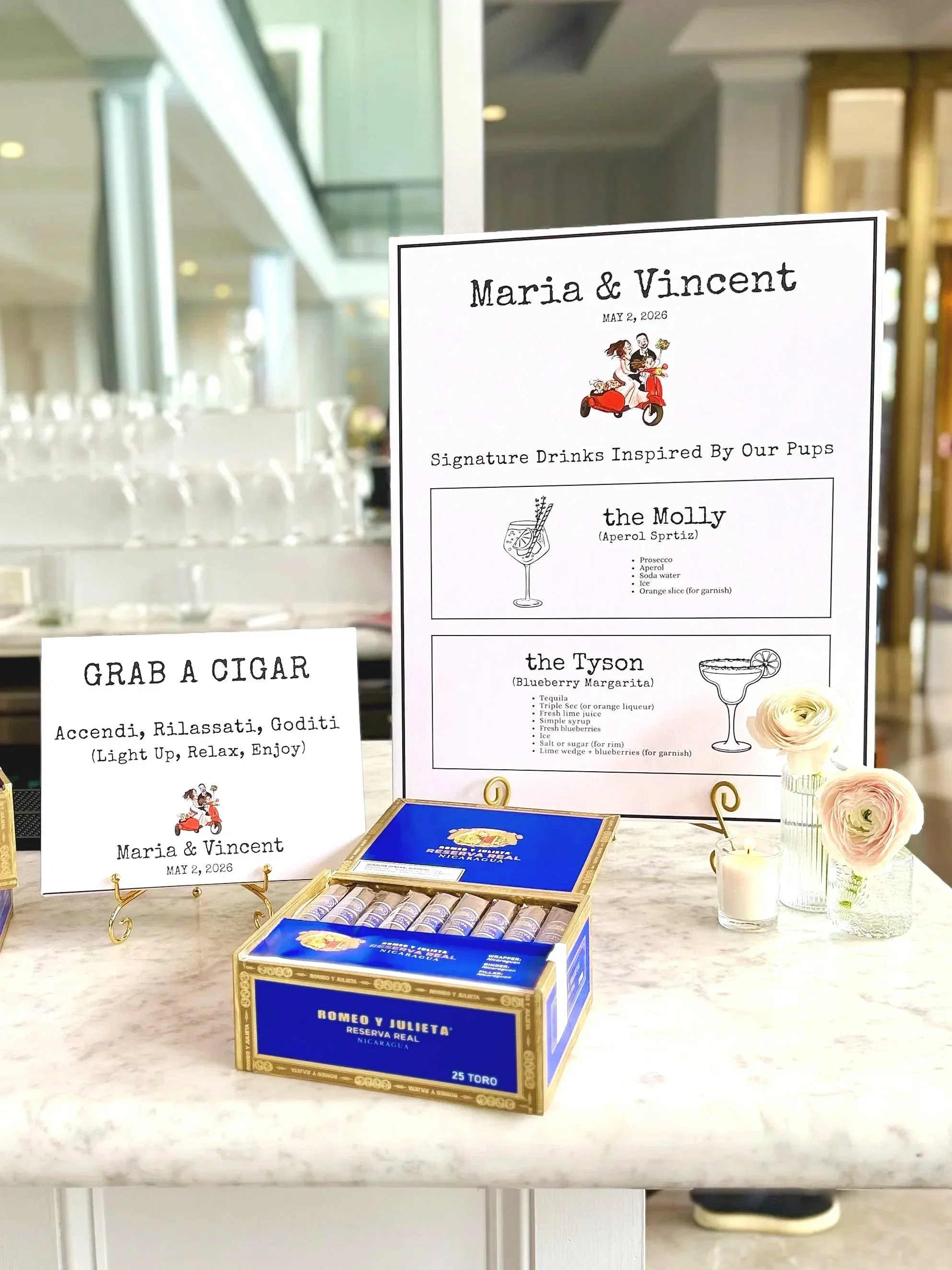 Wedding reception display with a sign featuring Maria and Vincent's names and wedding date, decorated with flowers and candles, and a box of Romeo y Julieta cigars on a marble counter.