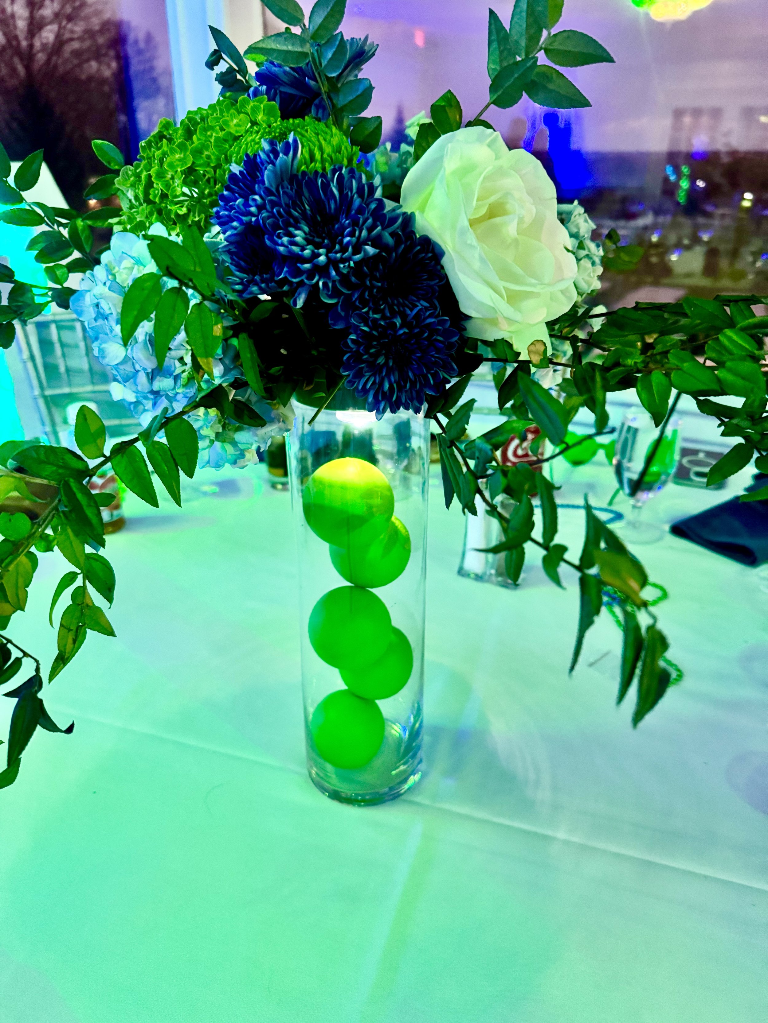 A floral centerpiece with white and blue flowers and green foliage in a tall glass vase filled with bright green balls, placed on a table.