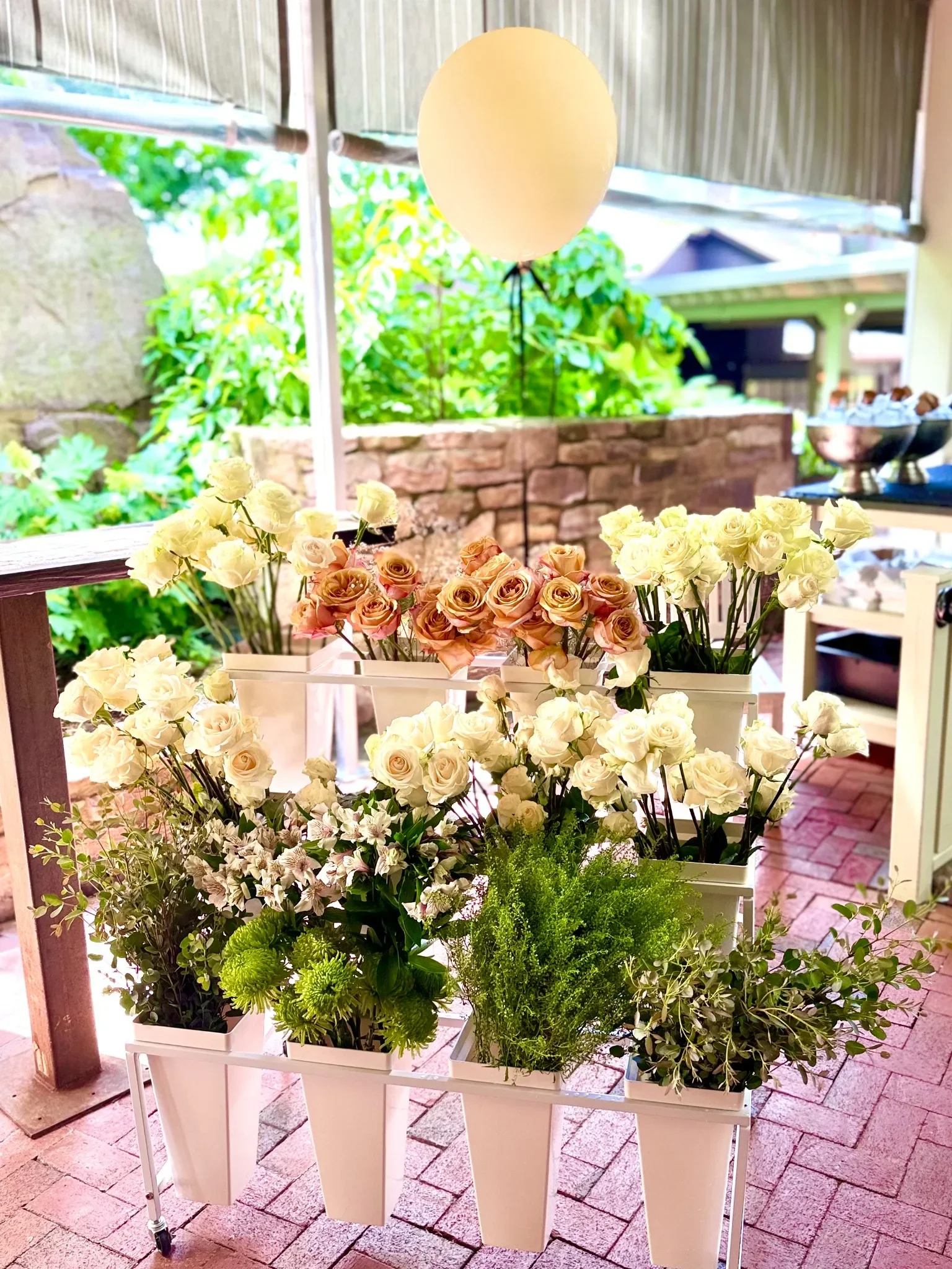 Flower display bloom bar with white and peach roses, green foliage, and an illuminated round lamp on a stand, set outdoors on a brick patio with a stone wall and greenery in the background.