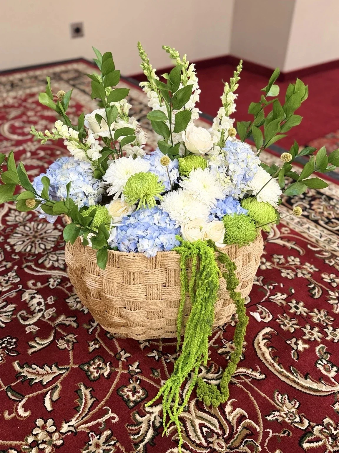 A basket with a woven pattern holding a colorful flower arrangement featuring white, light blue, green, and cream flowers, with green foliage, placed on a patterned red carpet.
