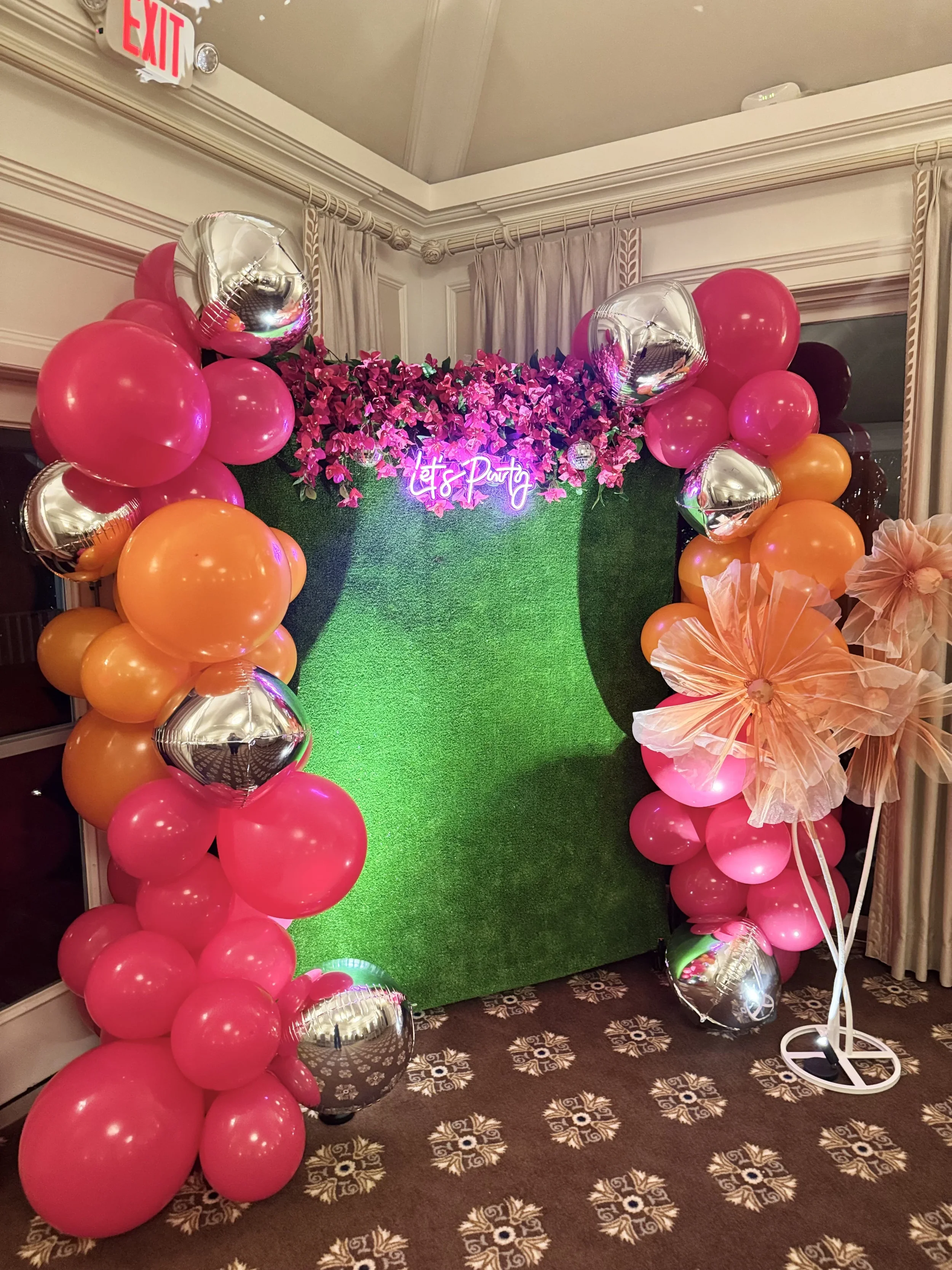 Decorative party backdrop with pink, orange, and silver balloons, pink artificial flowers, a neon sign reading "Let's Party," a green grass-like wall, and a flower stand with pink tissue paper flowers.