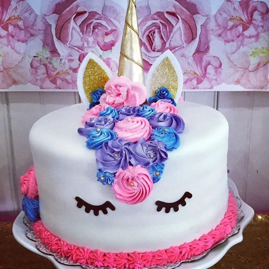 Unicorn-themed birthday cake with a golden horn, pink, purple, and blue frosting, and closed eyes, set against a pink floral background.
