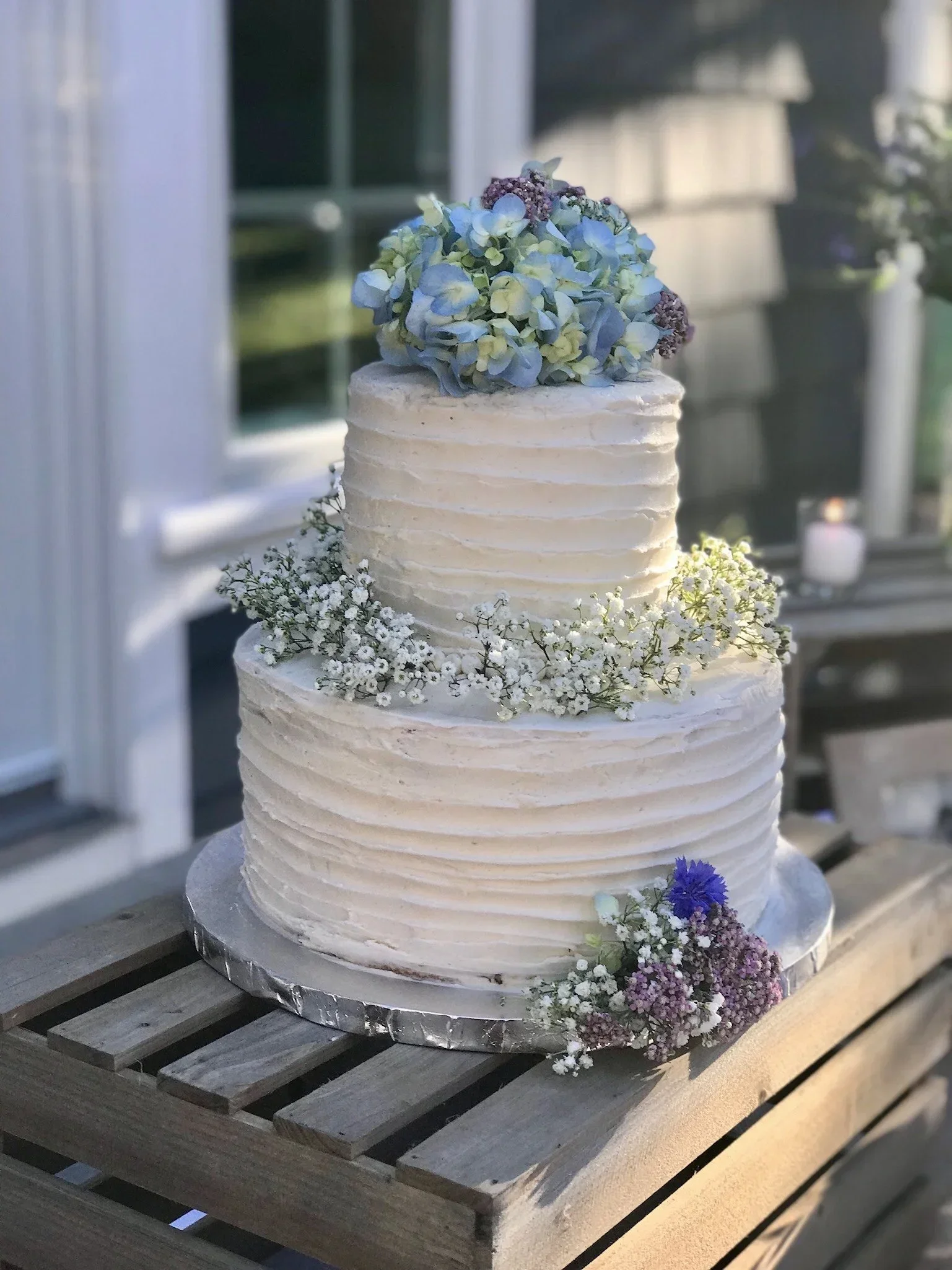 A three-tiered wedding cake decorated with light blue and purple flowers, displayed on a wooden surface.