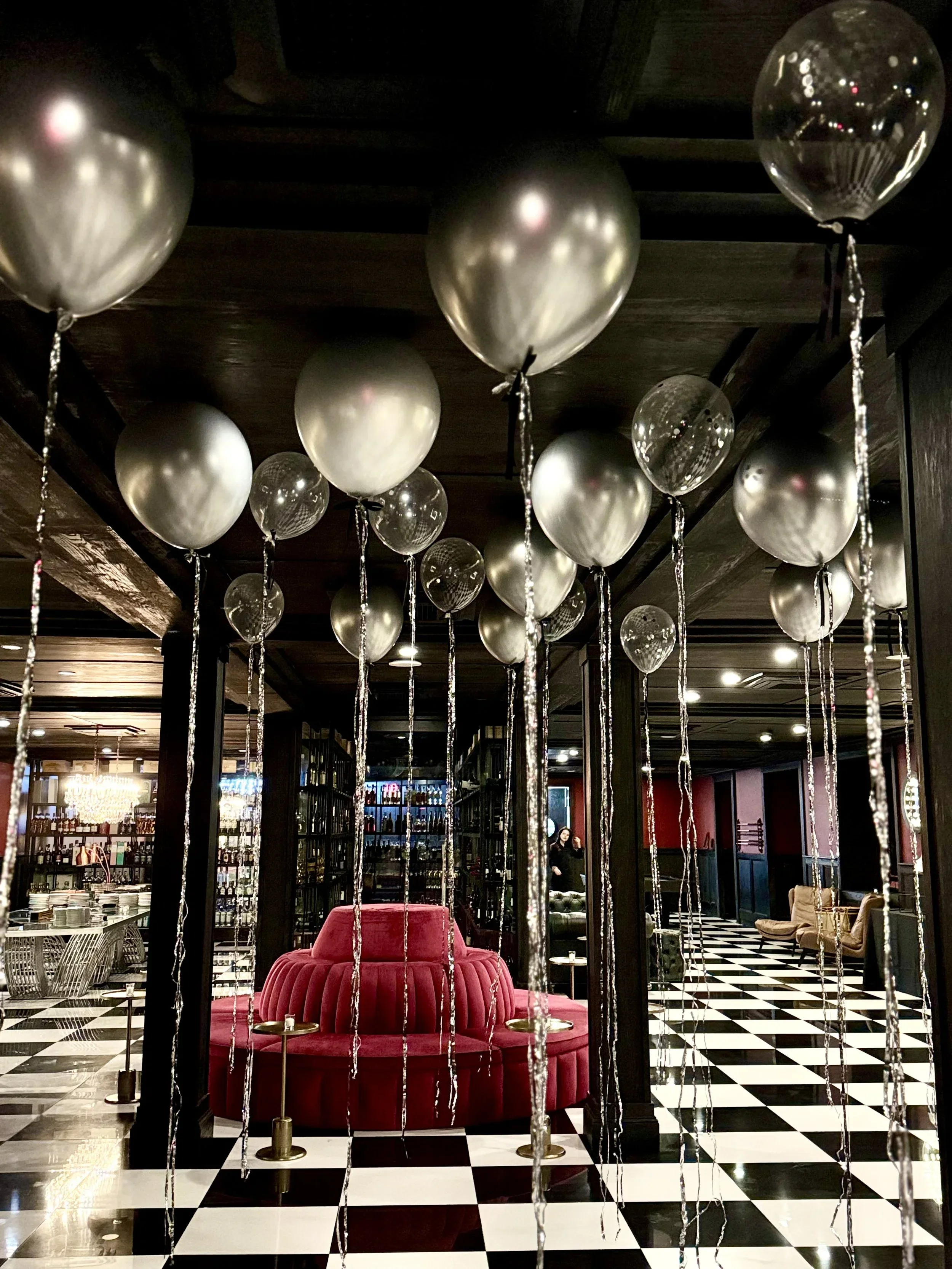 Interior of a decorated restaurant or bar with black and white checkered flooring, round balloons hanging from the ceiling, and plush seating including a red velvet booth.