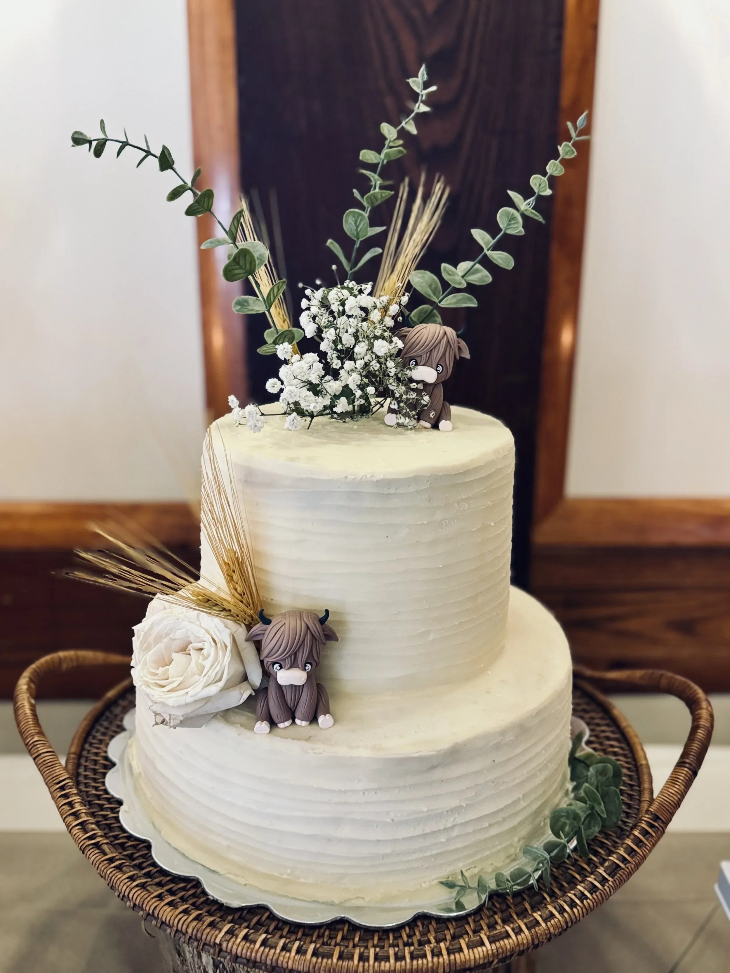A white, three-tiered wedding cake decorated with green eucalyptus leaves, white baby’s breath flowers, and a small figurine of a horse on the middle tier. The cake is on a silver platter, placed on a woven wicker table or stand.