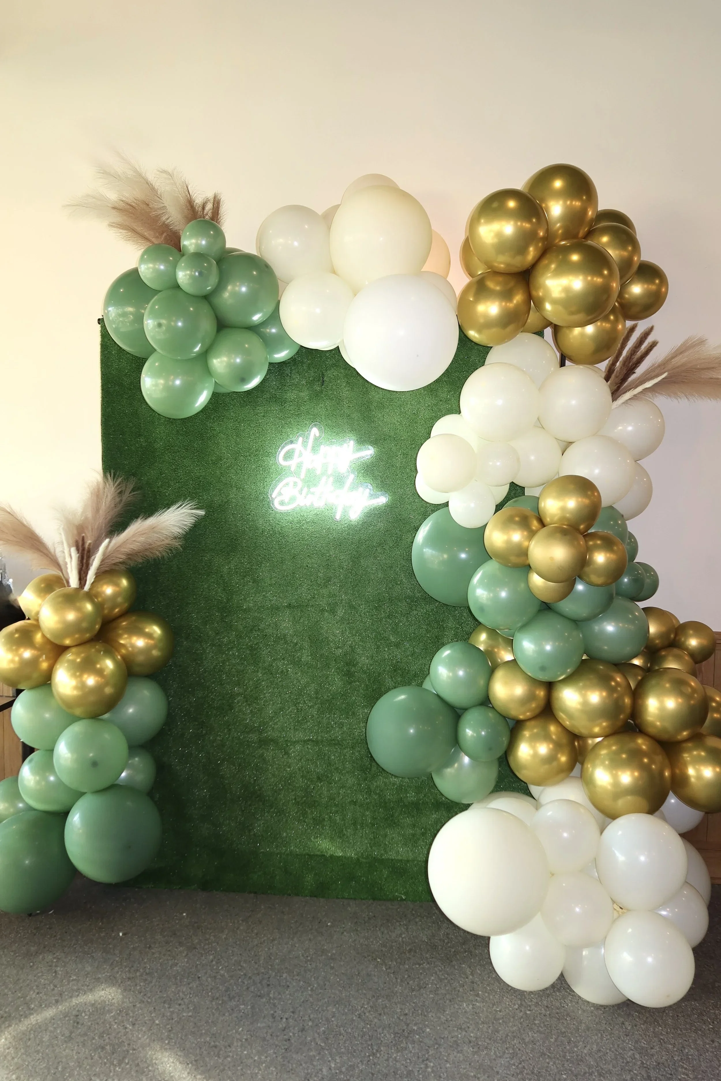 Decorative balloon arch with white, gold, and light green balloons, and pampas grass, with a neon sign reading 'Happy Birthday' on a green grass backdrop.
