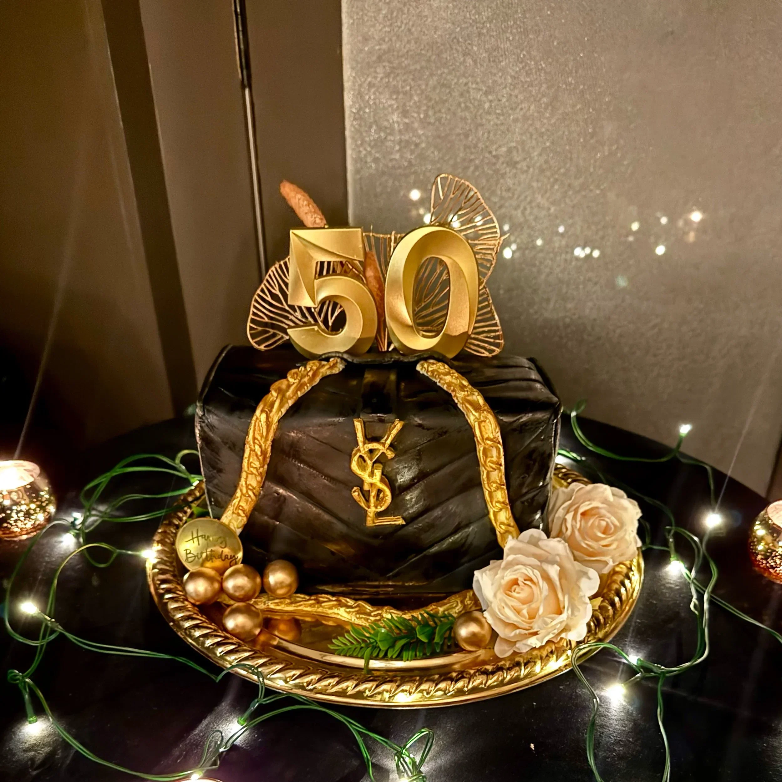 Decorative birthday display with large gold numbers '50', surrounded by gold and silver balls, cream roses, and green foliage on a black surface, with glittery black backdrop and string lights.