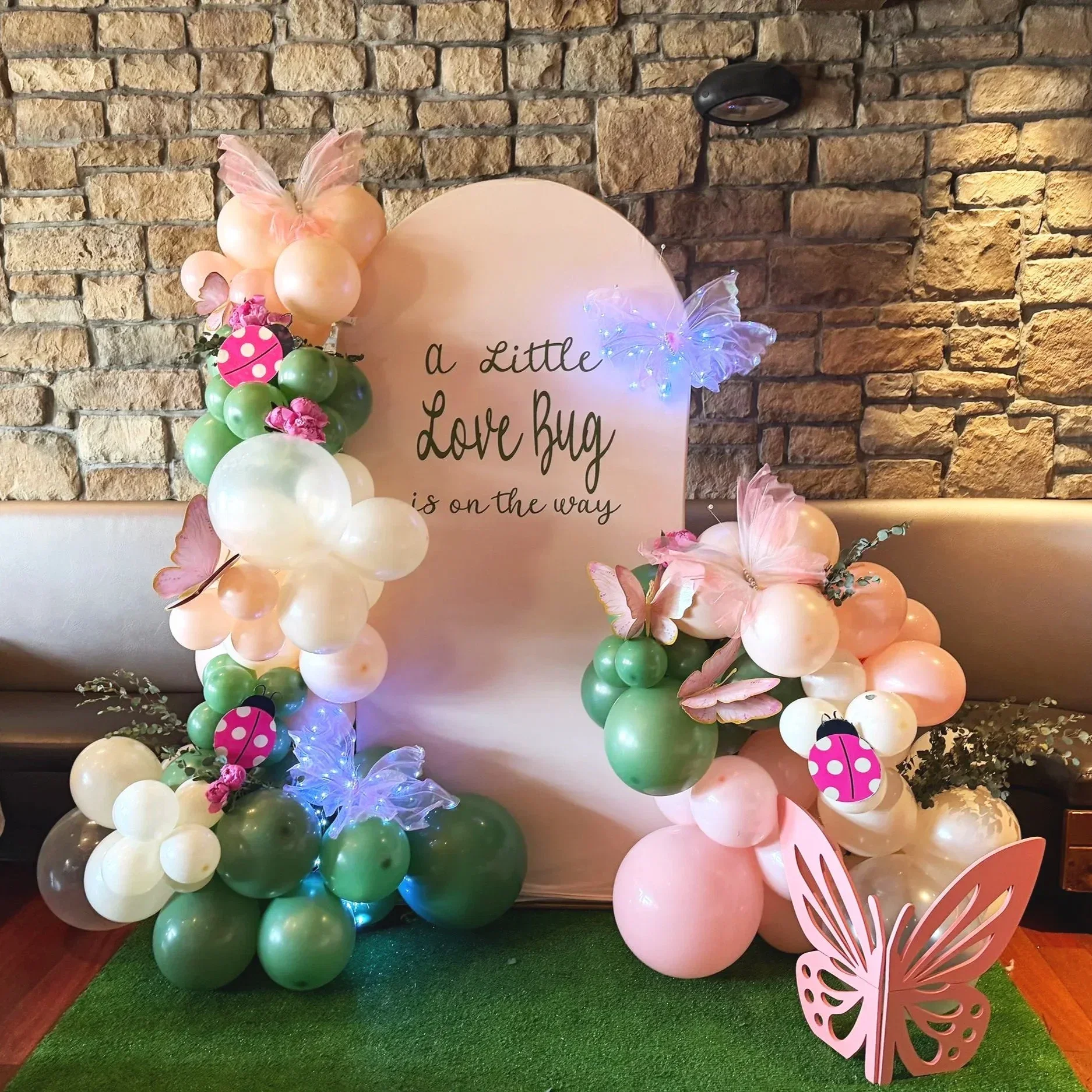 Decorative arrangement of pink, white, and green balloons with butterfly and ladybug accents, set against a brick wall with a pink circular backdrop containing text about a love bug.