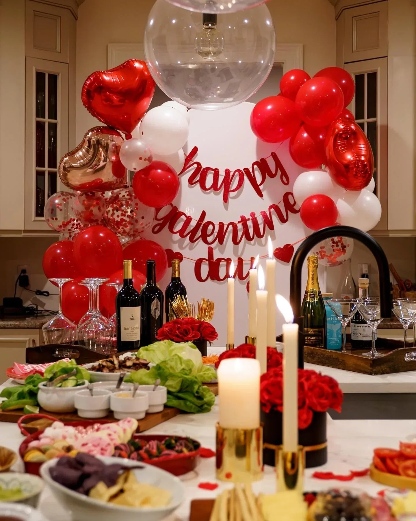 A kitchen decorated for Valentine's Day with red and white balloons, a "Happy Valentine's Day" banner, candles, and a table filled with food and drinks.