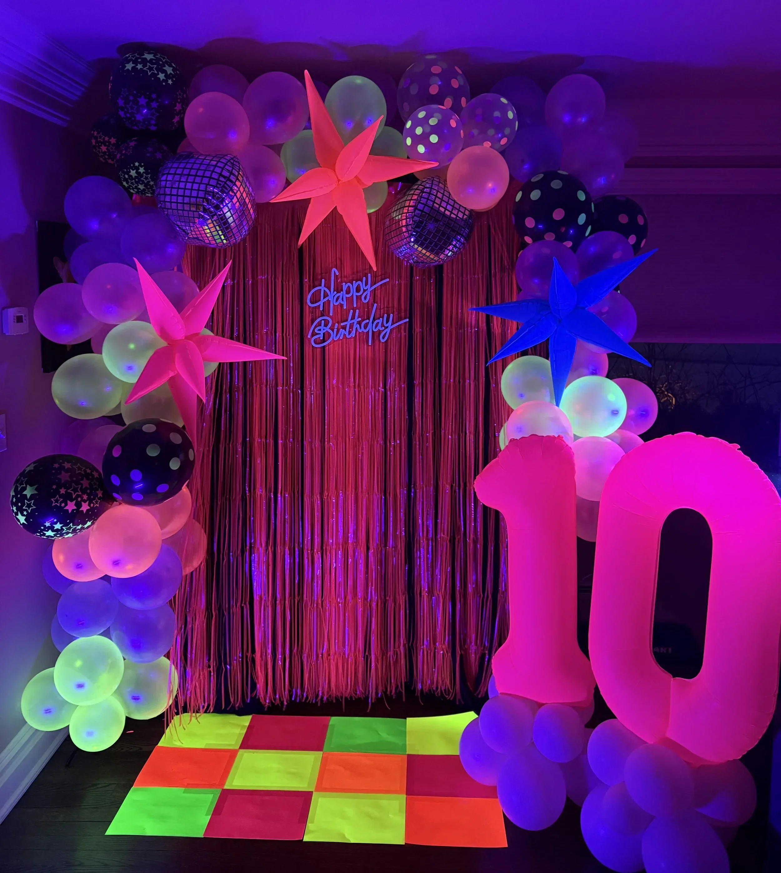 Neon birthday party decorations with balloons, star-shaped paper Lanterns, a reflective disco ball, a "Happy Birthday" sign, and a multicolored tiled floor.