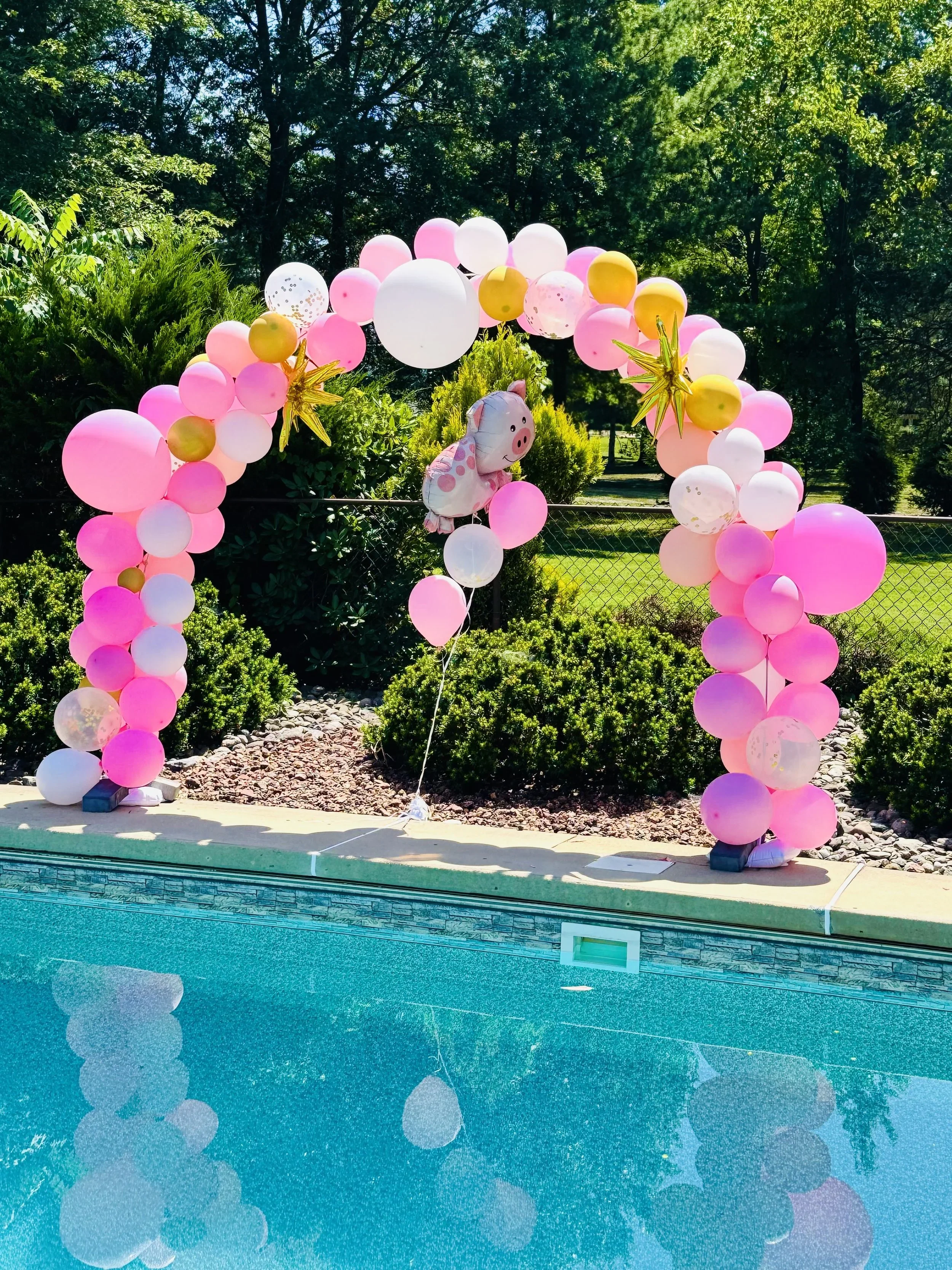 Decorative balloon arch near a swimming pool, with pink, white, and gold balloons, plush pig balloon, and gold star-shaped balloons, set outdoors against greenery and trees.