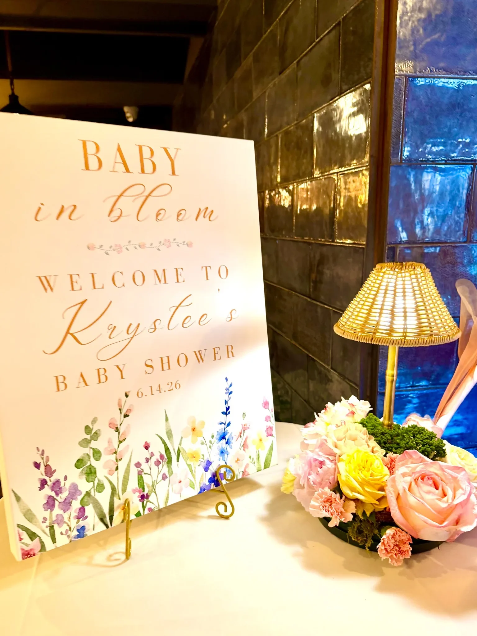 A floral-themed sign advertising a baby shower for Kraysee, with a bouquet of pink, yellow, and white flowers, a decorative gold lamp, and a black tiled wall in the background.