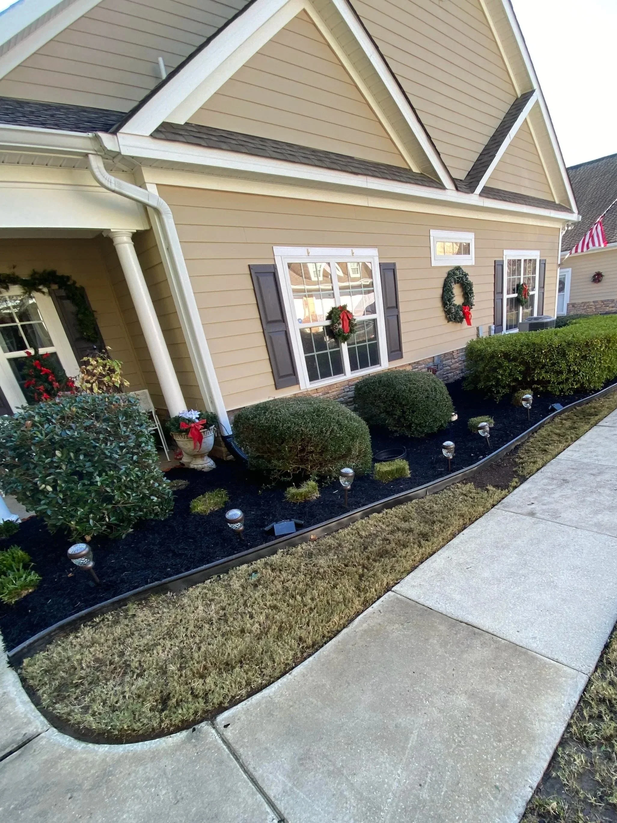 Front yard of a house decorated with Christmas wreaths and bows, with shrubs, a curved sidewalk, and outdoor lights.
