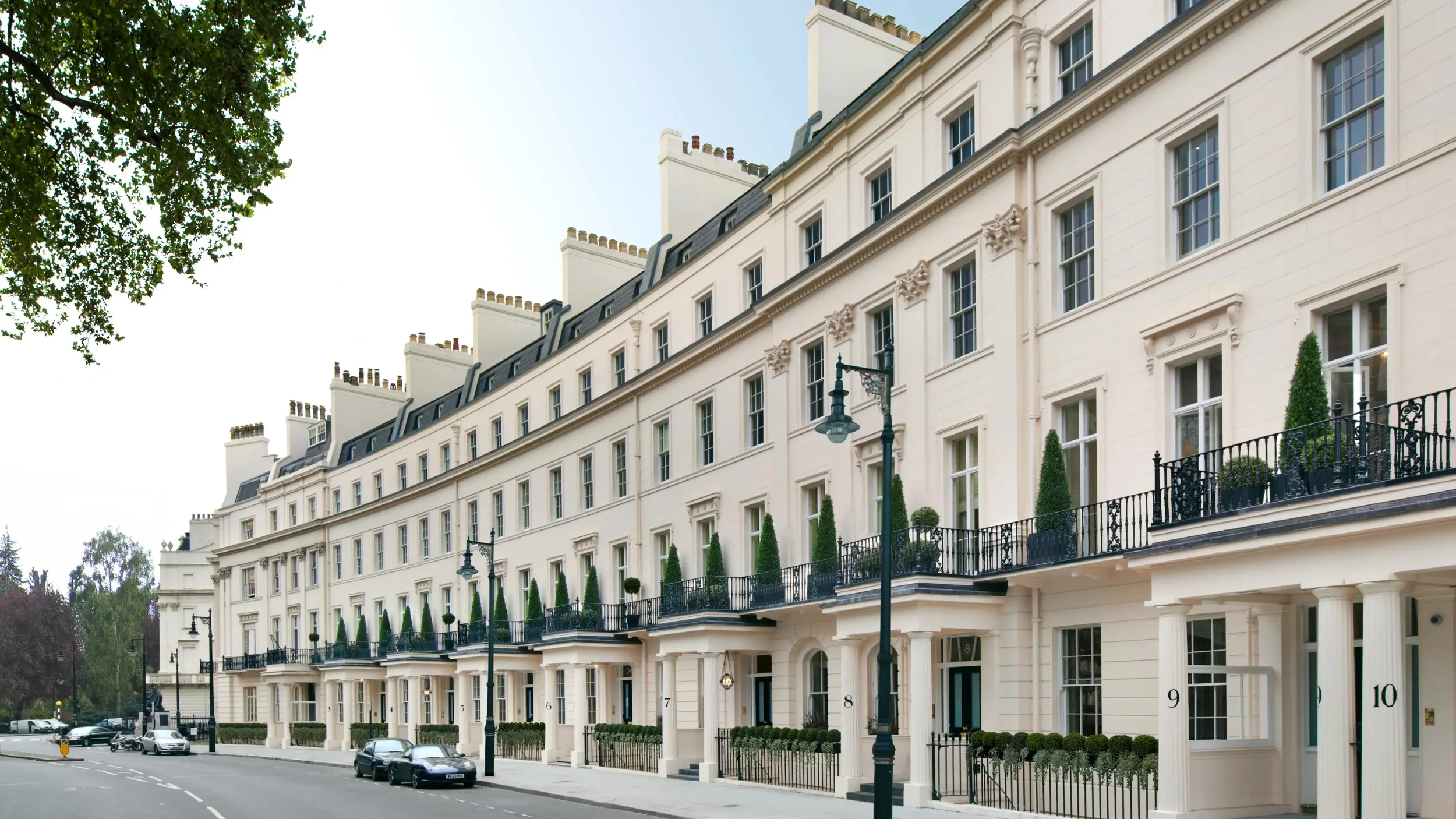 Row of elegant townhouses with white facades, black railings, potted plants, and parked cars along a street, with a large tree in the foreground.