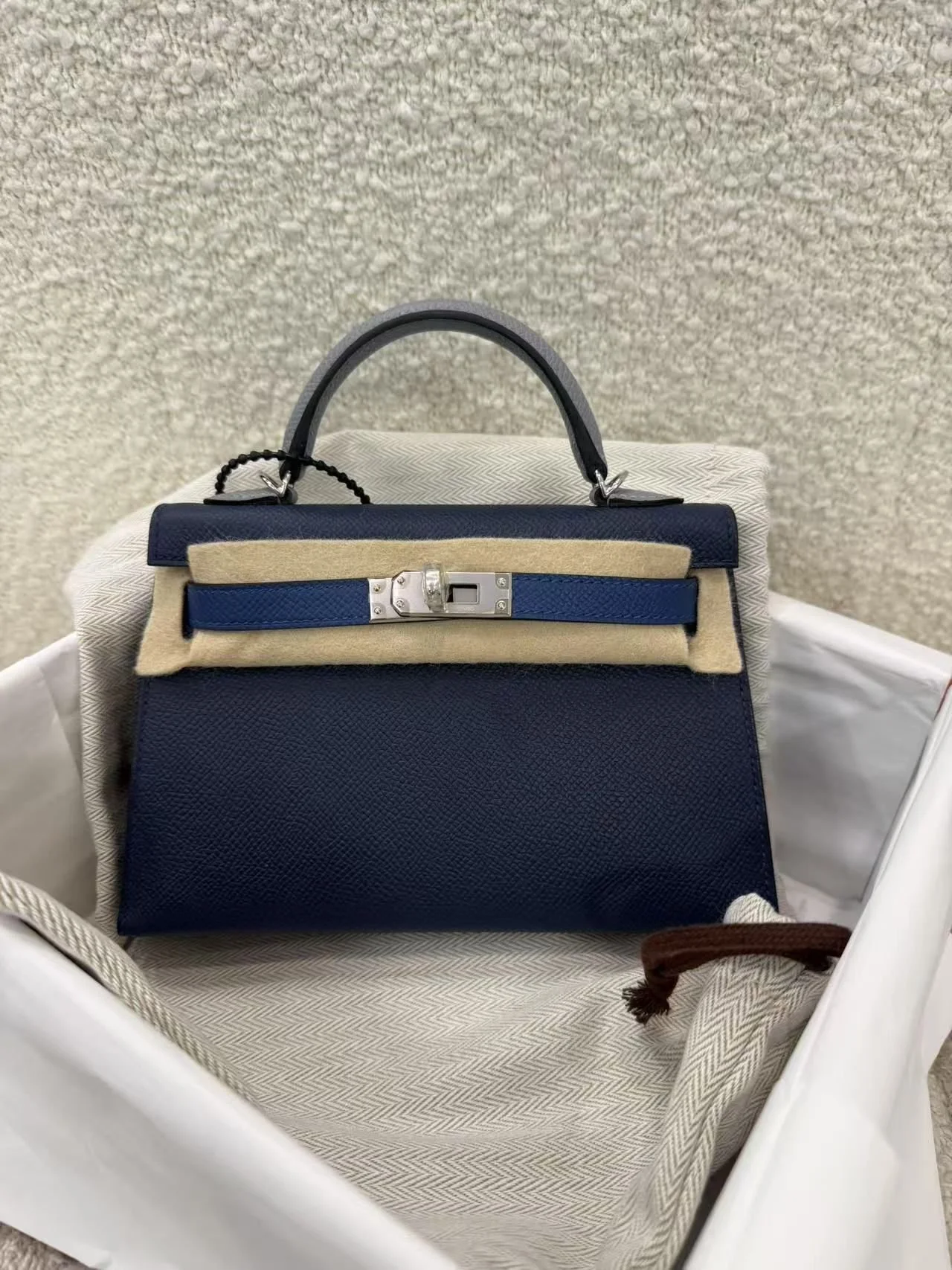 A navy blue designer handbag with a top handle, placed inside a white shopping bag lined with beige fabric, against a textured beige wall.