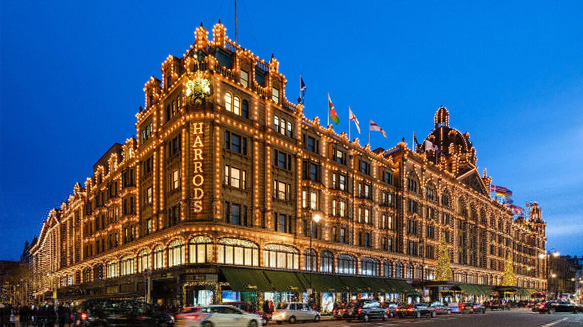 The Harrods department store illuminated with decorative lights in London at dusk, with flags on top and cars in front.