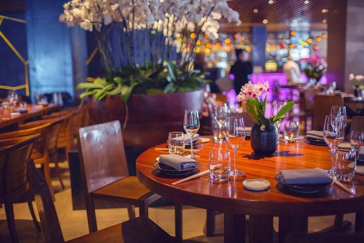 Set dining table with wine glasses, water glasses, chopsticks, napkins, and a black vase with white and purple flowers in a restaurant with warm lighting and purple accent lights, and large flower arrangements.