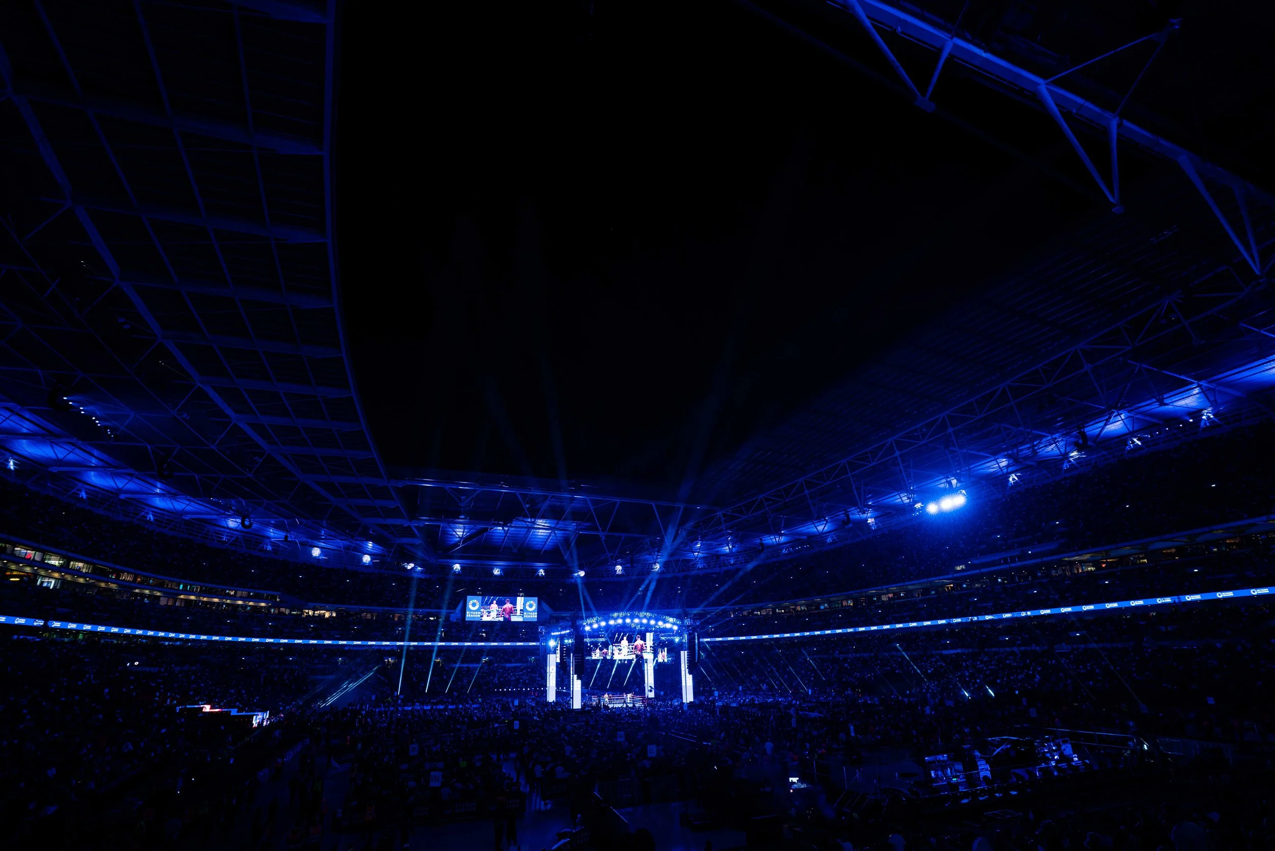 A large indoor stadium filled with spectators, illuminated with blue lights, hosting a sporting event on a central stage with screens.