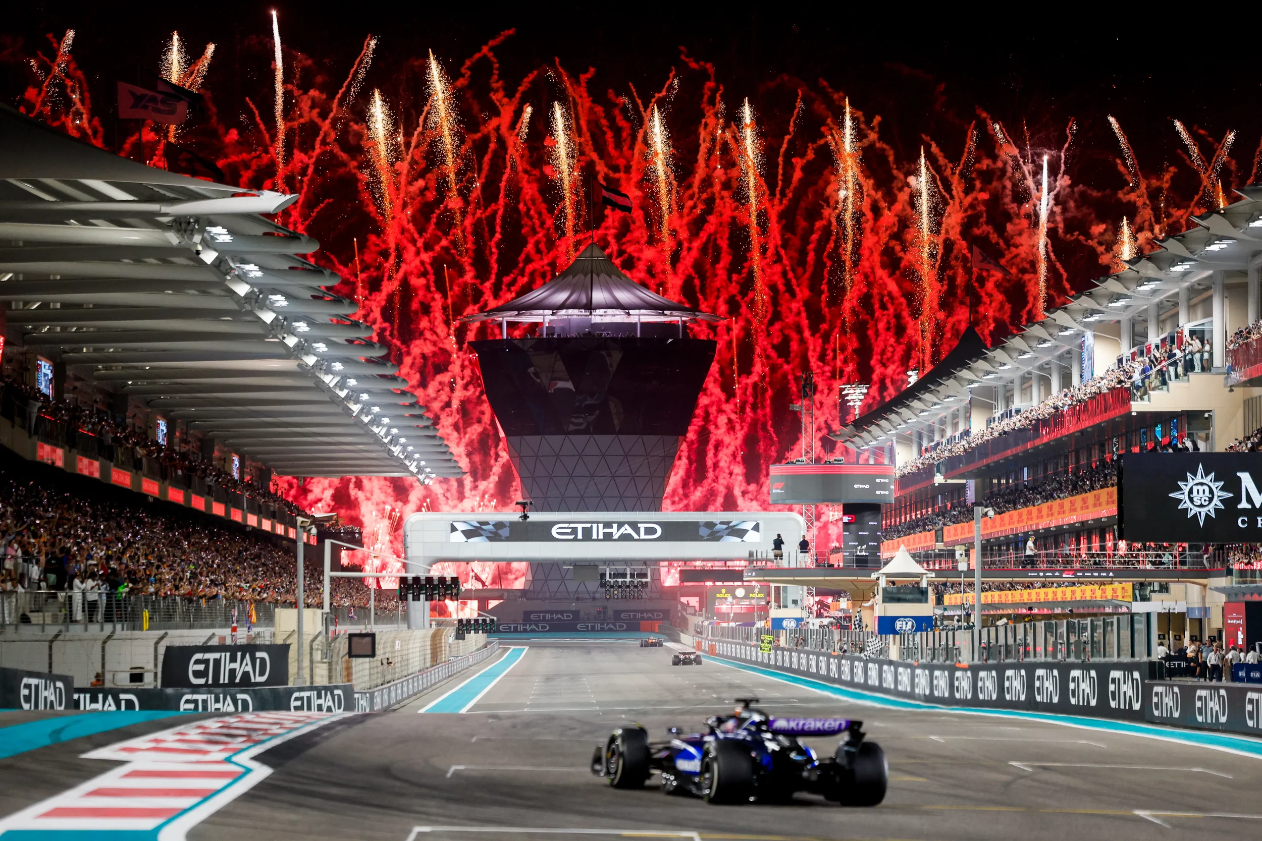 Race car driving on a racing track during an event at night, with fireworks in the sky and spectators in the grandstands.
