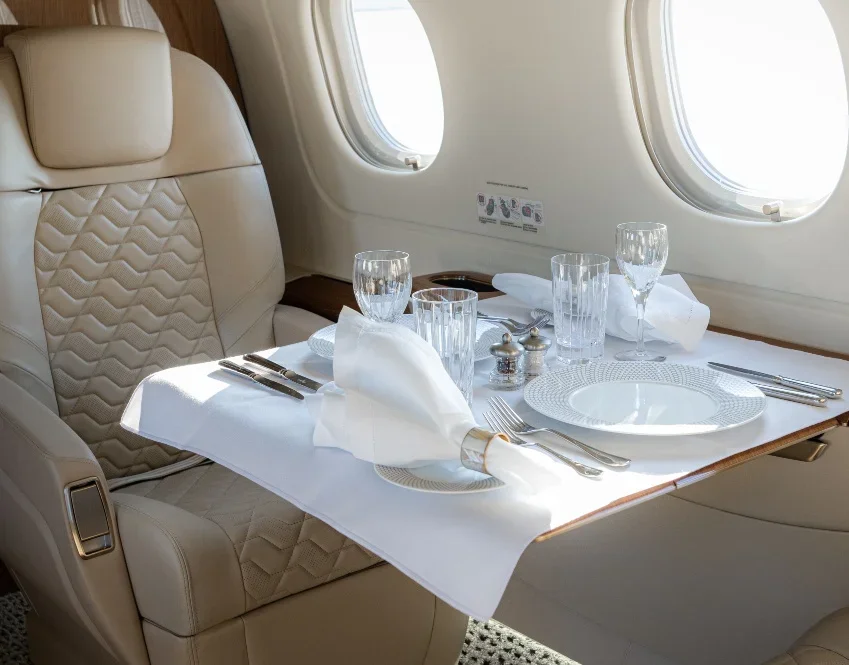 An airplane seat with a table set for dining, including white tablecloth, glassware, plates, napkins, and utensils, near airplane windows.