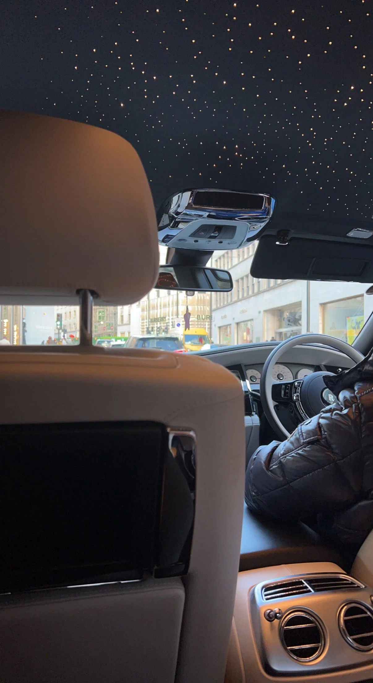 Inside a luxury car with a starry night sky ceiling design, showing the back of the front seats, the dashboard, and a street view through the windshield with buildings and vehicles.