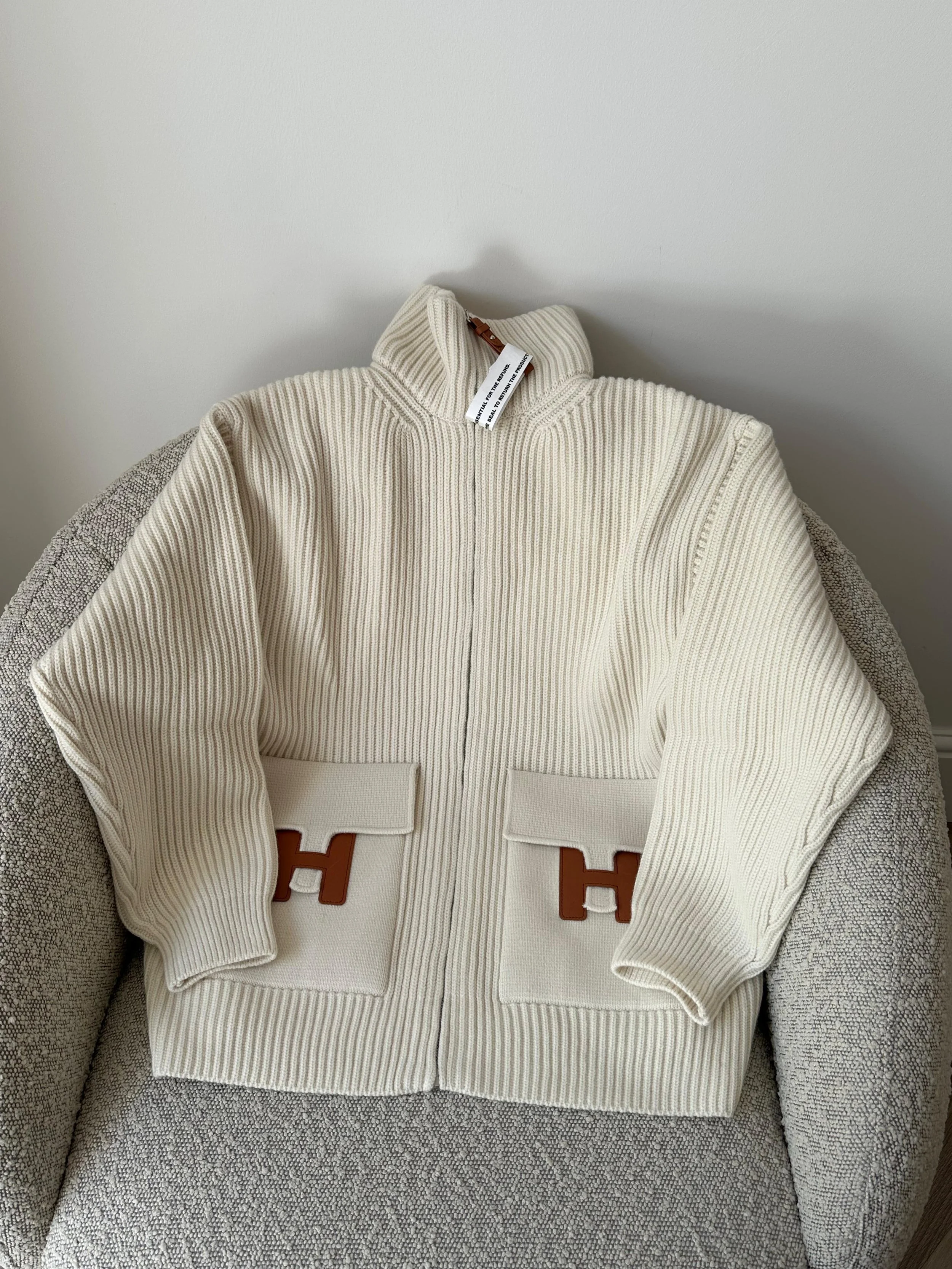 Cream-colored ribbed zip-up sweater with large front pockets, brown H logo patches, and a high collar, sitting on a light gray fabric chair.