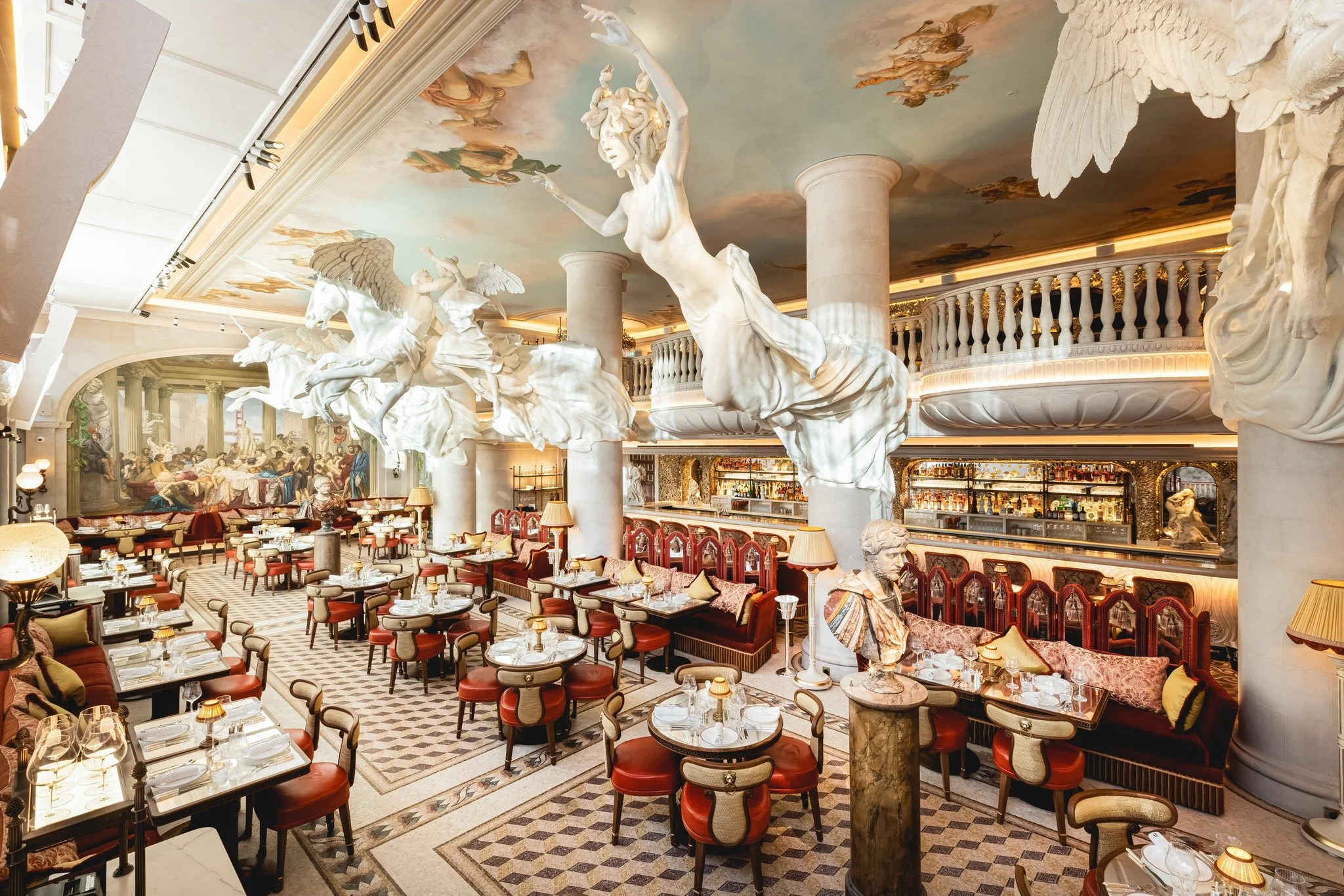 Elegant restaurant interior with classical sculpture decor, featuring large marble statues of angels and a reclining woman, intricately painted ceiling, patterned flooring, and neatly arranged tables with red chairs and white table settings.