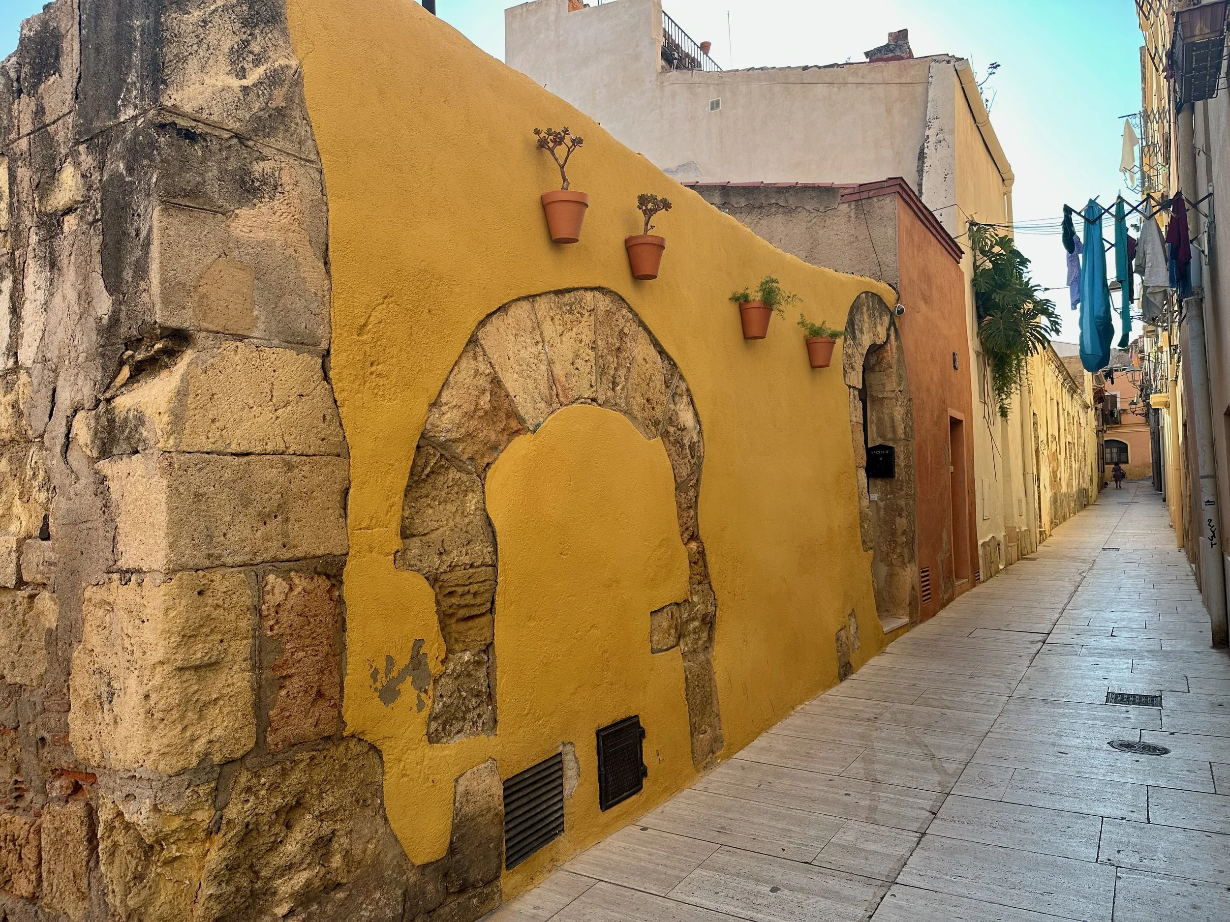 A narrow street in Tarragona, Spain with a yellow wall decorated with small potted plants, adjacent to stone and plaster buildings, with laundry hanging on a line to the right.