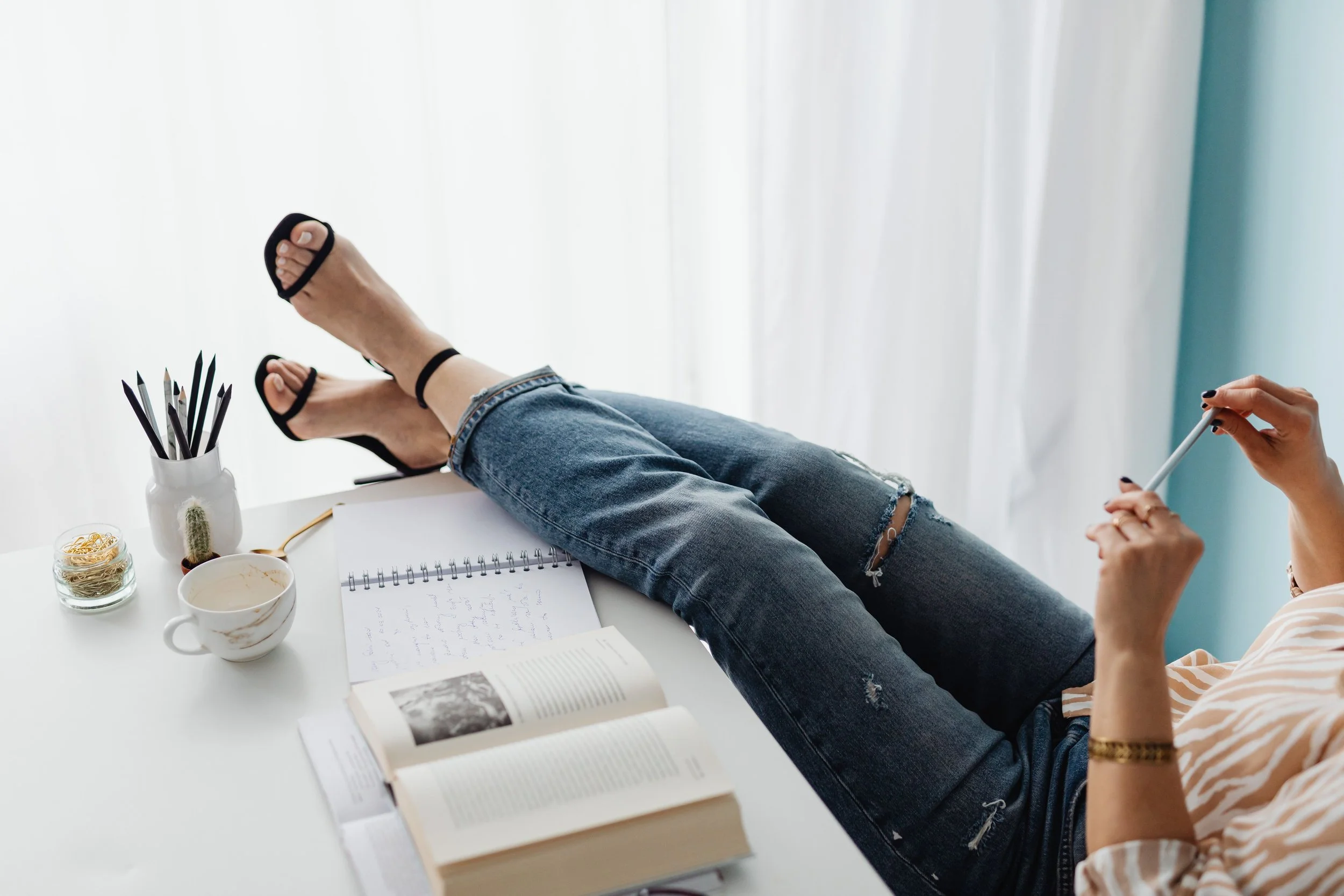 A person relaxing with legs crossed on a white table, wearing ripped jeans and black sandals, with a woman beside them holding a stylus. The table has an open book, a notebook with handwritten notes, a white cup with a beverage, a small glass jar of rubber bands, a small cactus in a white pot, and a container filled with black pencils.